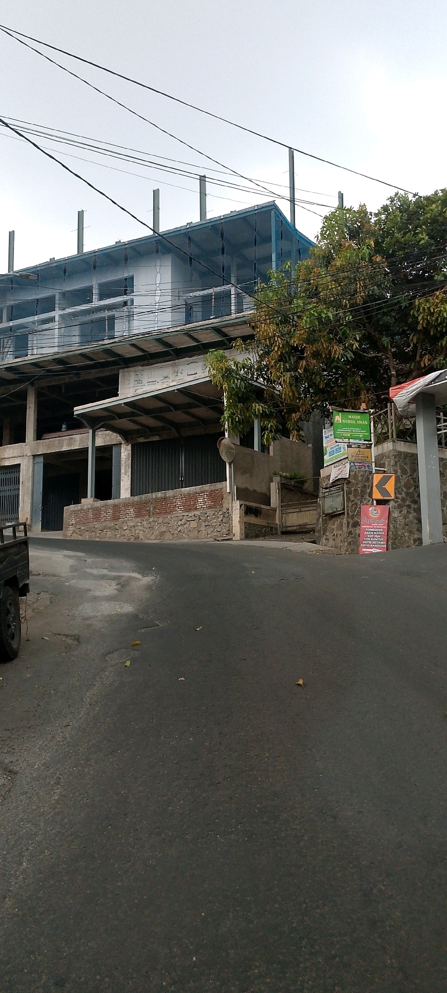 The image shows a multi-story building under construction, partially covered with blue netting, located on a sloping road in Wangunsari, Indonesia. The building appears to be a commercial or public structure, with exposed concrete pillars and metal framing on the upper levels, and a corrugated metal facade on the lower levels. A stone and brick wall runs along the front of the property. Several signs are posted on a stone pillar to the right of the building, including a green sign that reads "MASJID NURUL IMAN" indicating a mosque. A yellow directional sign points to the left. Trees with green and brown foliage are visible to the right of the building. A black pickup truck is parked on the left side of the road, partially in view. The road surface is asphalt and shows signs of wear and tear, with some potholes and scattered leaves. Overhead power lines crisscross the sky. The sky is overcast and appears to be a bright, diffused light. There are no people visible in the image.