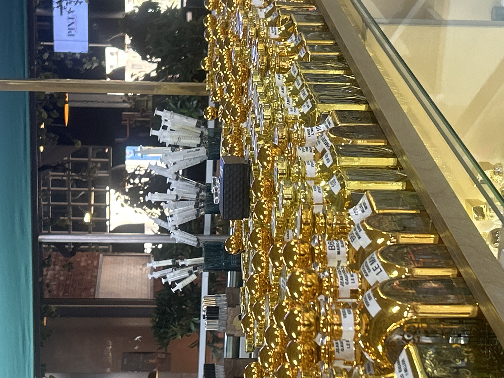 A well-lit, indoor retail environment displays rows of golden perfume bottles with ornate caps. The bottles are arranged on a multi-tiered shelf unit. In the background, several rows of syringes containing clear liquid are arranged in green cylindrical holders. Behind the syringes, a brick wall is visible, and a large window looks out onto an outdoor scene with greenery and a sign that reads "PINTA". The overall impression is that of a perfume store or boutique, potentially a high-end establishment given the presentation of the merchandise. The image does not provide specific visual cues to pinpoint the location within Almaty, Kazakhstan, beyond the context of an urban retail setting.