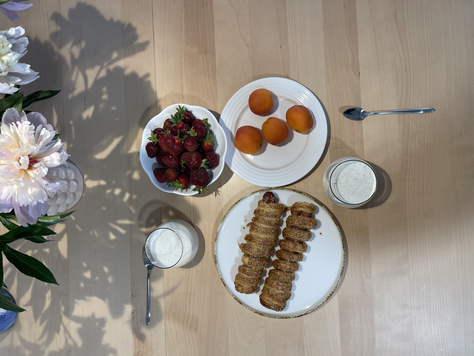 An overhead, top-down shot shows a breakfast setting on a light wood table. In the upper part of the frame, a vase holds several large, white and pink peonies. To the right of the vase is a bowl of ripe strawberries. Below the vase and to the left is a glass of white, creamy liquid, possibly yogurt or a smoothie, with a spoon resting on the table to its left. In the lower left of the frame is a white plate with two pastries that appear to be sausages wrapped in dough and sprinkled with sesame seeds. To the right of that is another white plate holding four ripe apricots. Below that plate is a single spoon, and to the left of the apricots is another glass of white, creamy liquid.