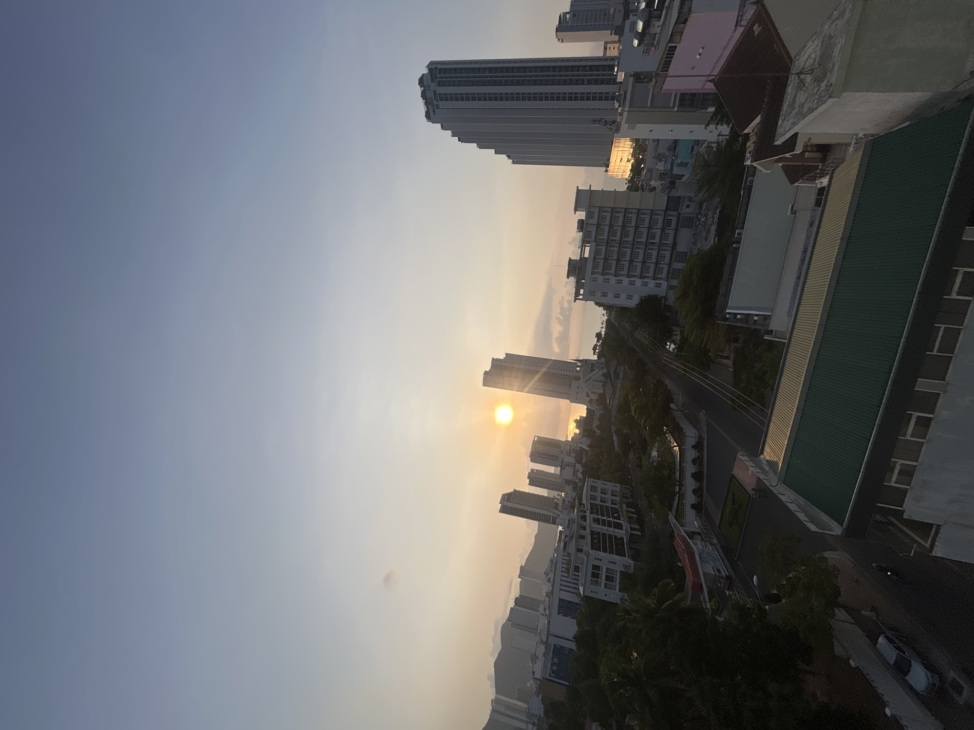 The image captures a view of the city of Bac Nha Trang Ward, Vietnam, during what appears to be either sunrise or sunset, given the position of the sun in the sky. High-rise buildings dominate the urban landscape, with several skyscrapers visible. The sky exhibits a gradient of colors from a light blue to an orange hue near the horizon where the sun is positioned. A street is partially visible, lined with greenery. Some rooftops are also visible.