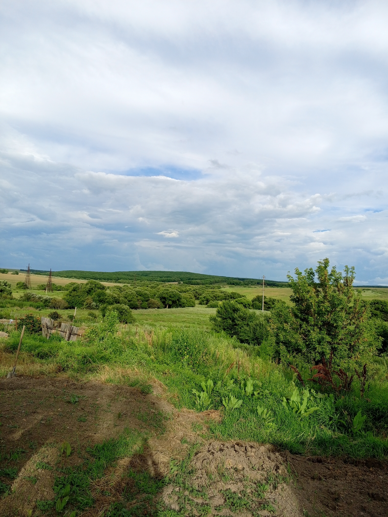 The image captures a rural landscape under a cloudy sky. In the foreground, a patch of tilled soil transitions into overgrown green grass. A wooden fence, partially broken, stands to the left, with a shovel leaning against it. Beyond this, rolling green fields are dotted with clusters of trees and bushes. In the distance, hills form the horizon, and power lines with tall poles are visible, indicating a rural or semi-rural setting. The sky is filled with dramatic, heavy clouds, suggesting the possibility of rain. The scene is devoid of people.