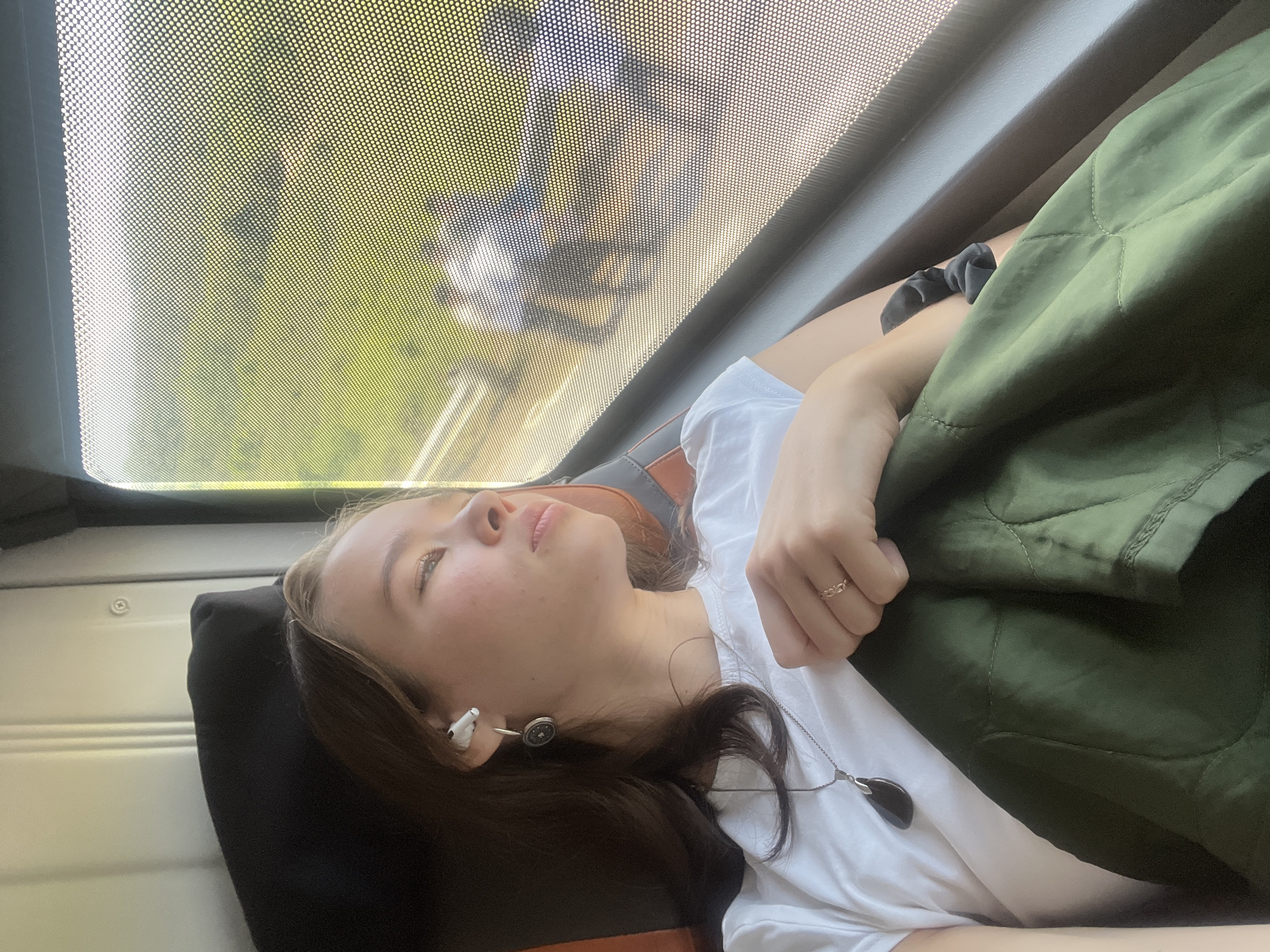 A woman is lying down, possibly on a train or bus, near a window with a dotted screen. She is resting her head on a dark pillow and has on a white shirt and a green quilted jacket is partially covering her. She has headphones in her ear and is wearing a necklace with a dark pendant and a ring on her finger. The background visible through the window appears to be greenery. There is no discernible evidence to determine the specific location in Khánh Vĩnh, Vietnam.