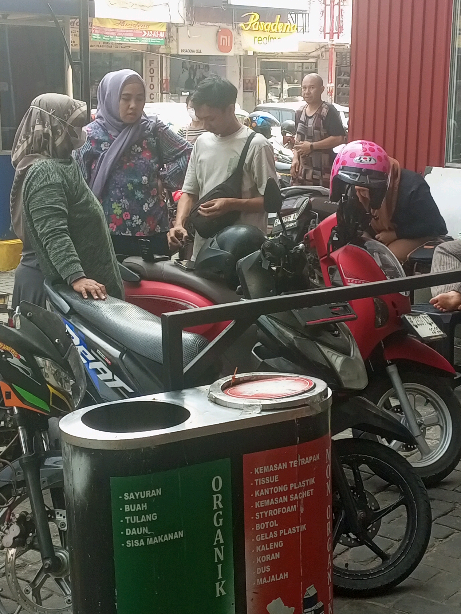 The image depicts a street scene in Bandar Lampung, Indonesia, with a group of people gathered around motorcycles and a recycling bin. In the foreground, a black and silver recycling bin is visible, divided into sections for organic and inorganic waste. The organic section, colored green, lists types of organic waste in Indonesian, such as "SAYURAN" (vegetables), "BUAH" (fruits), "TULANG" (bones), "DAUN" (leaves), and "SISA MAKANAN" (food scraps). The inorganic section, colored red, also lists types of inorganic waste in Indonesian, including "KEMASAN TETRAPAK" (Tetra Pak packaging), "TISSUE" (tissue), "KANTONG PLASTIK" (plastic bags), "KEMASAN SACHET" (sachet packaging), "STYROFOAM," "BOTOL" (bottles), "GELAS PLASTIK" (plastic cups), "KALENG" (cans), "KORAN" (newspapers), "DUS" (cardboard boxes), and "MAJALAH" (magazines).
Several motorcycles are parked in the scene, including a red scooter in the mid-ground and a black scooter in the foreground. A person wearing a pink helmet with polka dots is seated on the red scooter, while another person in a dark blue shirt is partially visible behind them. To the left, a person wearing a green long-sleeved shirt and a headscarf stands next to a motorcycle, their hand resting on the seat. Another person wearing a light purple hijab and a floral patterned top is standing in the center, looking towards the right. A man with a shaved head and wearing a patterned vest over a dark shirt stands behind the people in the center.
The background shows a street with buildings, signage, and parked vehicles, suggesting an urban environment. One sign reads "Pasadena" in yellow letters with Indonesian text below. The overall atmosphere appears to be a casual street interaction or a brief stop in a commercial area.