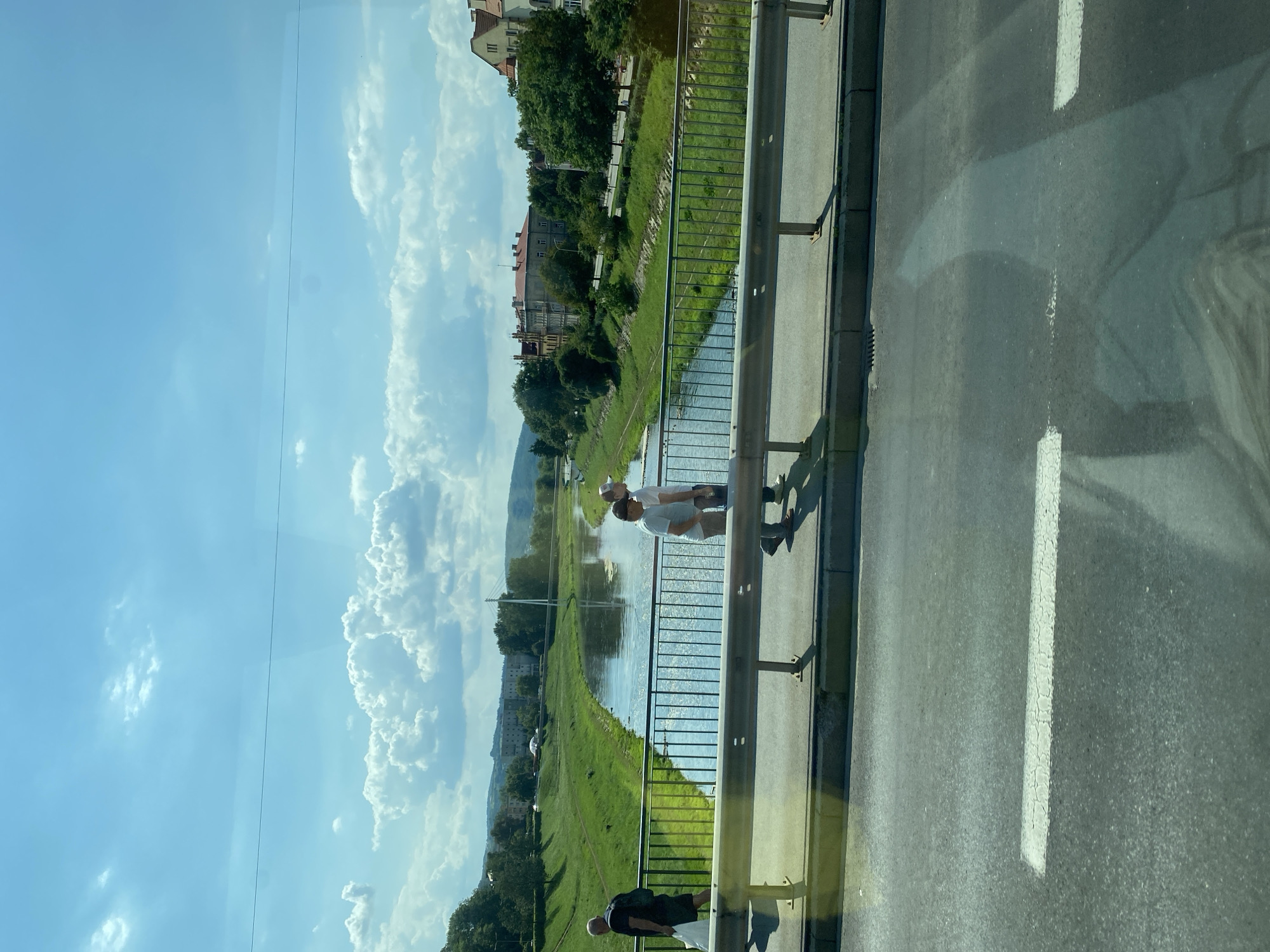 The image depicts a view from a bridge looking across a river in Przemyśl, Poland. On the bridge, three individuals are visible. To the left, a man wearing a black t-shirt and shorts walks away from the camera, carrying a white plastic bag. In the center, two men, both wearing white t-shirts and shorts, walk side-by-side, facing away from the camera. The man on the left has a baseball cap on.
The bridge has a metal railing along its edge. Beyond the railing, a river flows, reflecting the sky. The riverbanks are lined with lush green grass. On the far side of the river, buildings with red-tiled roofs and trees are visible, suggesting an urban or town setting. In the distance, hills or mountains form the backdrop, and a white suspension bridge can be seen. The sky is blue with scattered white, fluffy clouds. The foreground shows a section of asphalt road with white lane markings. The overall scene suggests a sunny day with a pleasant atmosphere.