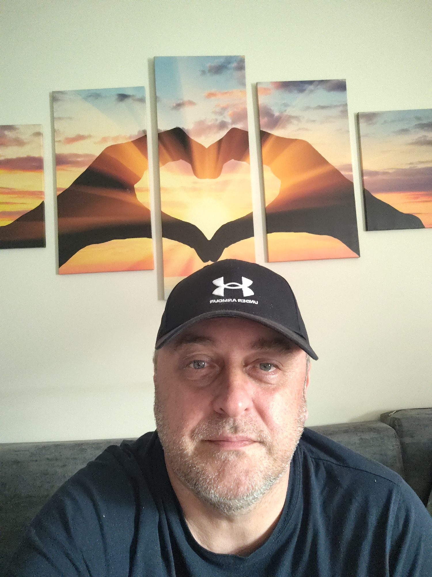 A middle-aged man with a fair complexion, light stubble on his face, and light-colored eyes is captured in a front-facing photograph, likely a selfie. He wears a dark blue or black t-shirt and a black baseball cap with the white, mirrored "Under Armour" text and logo visible on the front. His expression is neutral as he looks directly at the camera.
He is seated indoors, likely in a living room, on a dark gray fabric couch that is partially visible in the lower portion of the image. The wall behind him is a plain, light color, possibly off-white or very light green. Dominating the background is a five-panel wall art piece depicting a sunset or sunrise scene. The central three panels feature two silhouetted hands forming a heart shape, with the sun radiating light from within the heart. The surrounding panels complete the scenic view of a warm sky with orange, yellow, and red hues blending into a blue, cloudy sky. The overall impression suggests a relaxed, indoor setting in Huntingdonshire, United Kingdom.