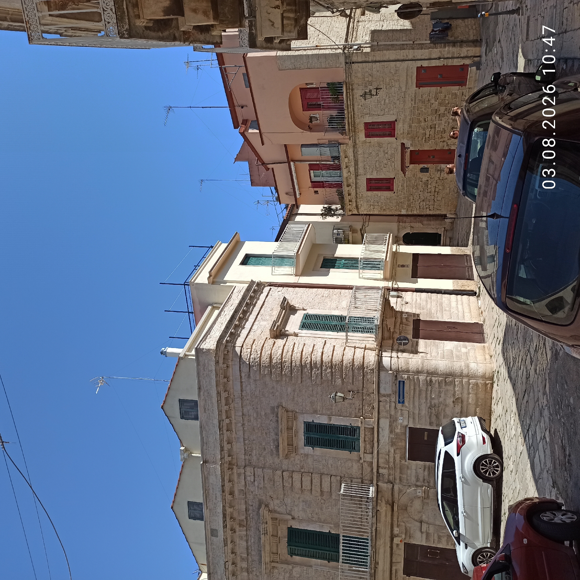 The image depicts a street scene in Ruvo di Puglia, Italy, on a clear day. The architecture features light-colored stone buildings with shuttered windows and balconies. In the foreground, a dark-colored car is parked on a cobblestone street, with its rear visible. To the left, a white SUV is parked. Further back, a multi-story building with a pink facade and arched balconies is visible. People are seen on a balcony of this pink building, appearing to be interacting. Another building to the left is constructed of light stone with prominent horizontal banding and features dark wooden doors. Antennas are visible on the rooftops of several buildings. The scene is illuminated by bright sunlight, casting shadows on the ground. A street sign with Italian text is mounted on the wall of one of the buildings. The timestamp "03.08.2026 10:47" is superimposed on the image.