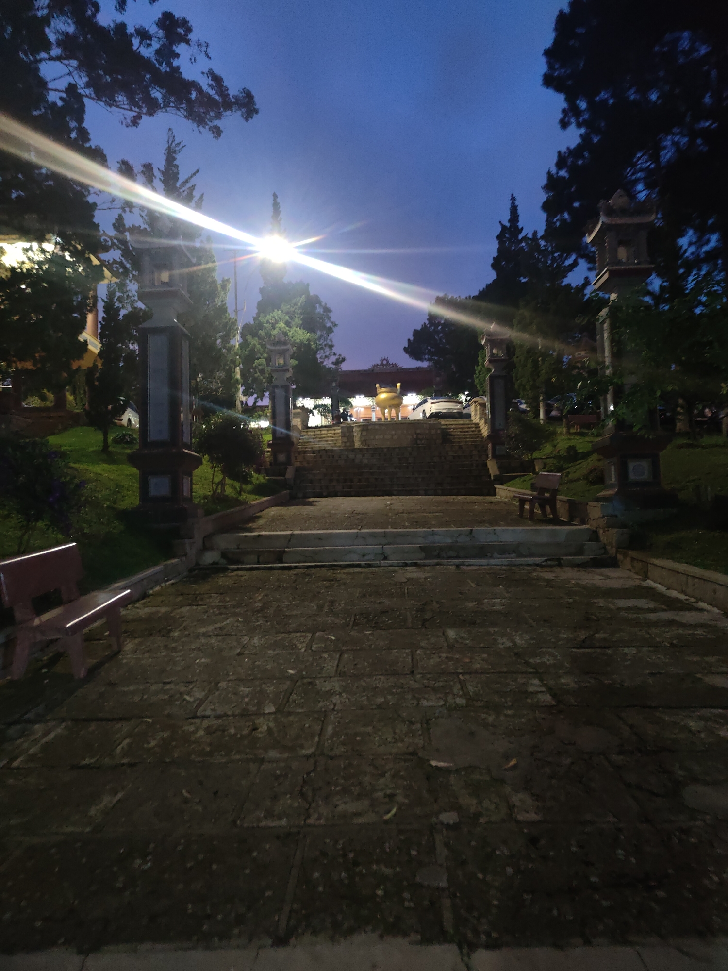 This image captures an outdoor scene at dusk in Da Lat, Vietnam. The foreground is dominated by a stone pathway, appearing somewhat damp and speckled with debris. To the left, a dark red bench is visible. The pathway leads upwards towards a wide, multi-tiered stone staircase.
Flanking the staircase are two tall, ornate stone pillars topped with lantern-like structures. These pillars are positioned at intervals along the ascent, creating a grand entrance. Beyond the top of the stairs, a building with a red-tiled roof is partially visible, illuminated by interior lights. In front of this building, a large, golden urn or pot sits prominently. A white car is parked near the building.
The background is filled with lush green trees, some with dark, dense foliage that contrasts with the lighter green of the grass on the sloping ground beside the stairs. The sky above is a deep twilight blue, suggesting the time is shortly after sunset. A bright light source, possibly a streetlamp, creates a strong lens flare effect, radiating across the upper left portion of the image. The overall atmosphere is serene and somewhat mystical, evoked by the dim lighting, the architectural elements, and the natural surroundings. There is no visible text in the image.