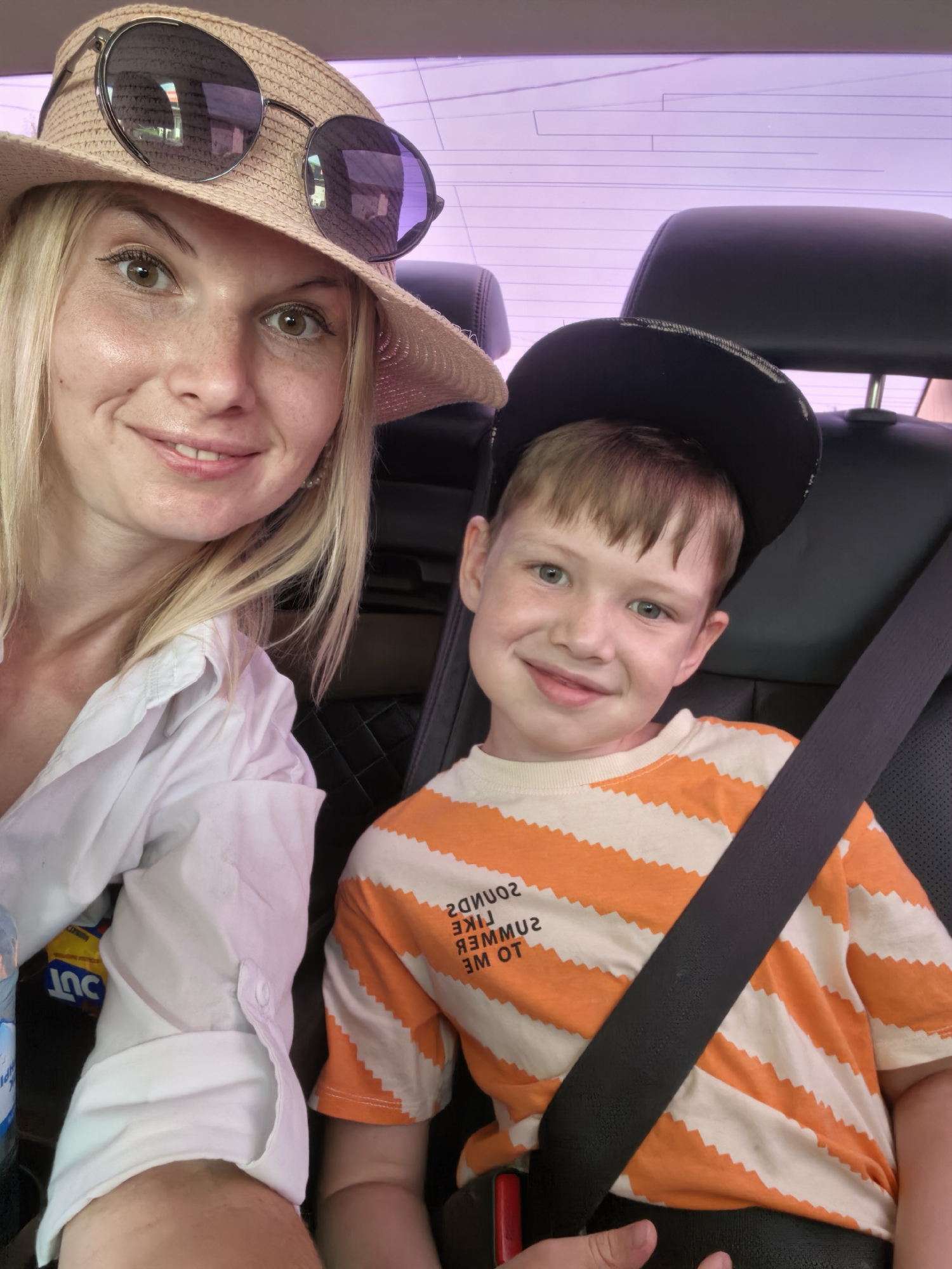 In this media file, a woman and a young boy are seated in the back of a vehicle. The woman, wearing a straw hat and sunglasses, is positioned on the left side of the frame. She has blonde hair and is smiling towards the camera. She is wearing a white shirt. The boy is on the right, also facing the camera, and wearing a black baseball cap and an orange and white striped t-shirt. He has a slight smile. Both are wearing seat belts, indicating they are on a journey. The background shows the interior of a car, with headrests and the window tinting the scene with a soft purple hue, suggesting it might be daytime or a lightly overcast day. The overall mood appears relaxed and happy, as both subjects are smiling. There is some text visible on the boy's shirt, but it is partially obscured and reversed, making it difficult to read clearly. The context provided places the scene in Sterlitamak, Russia.