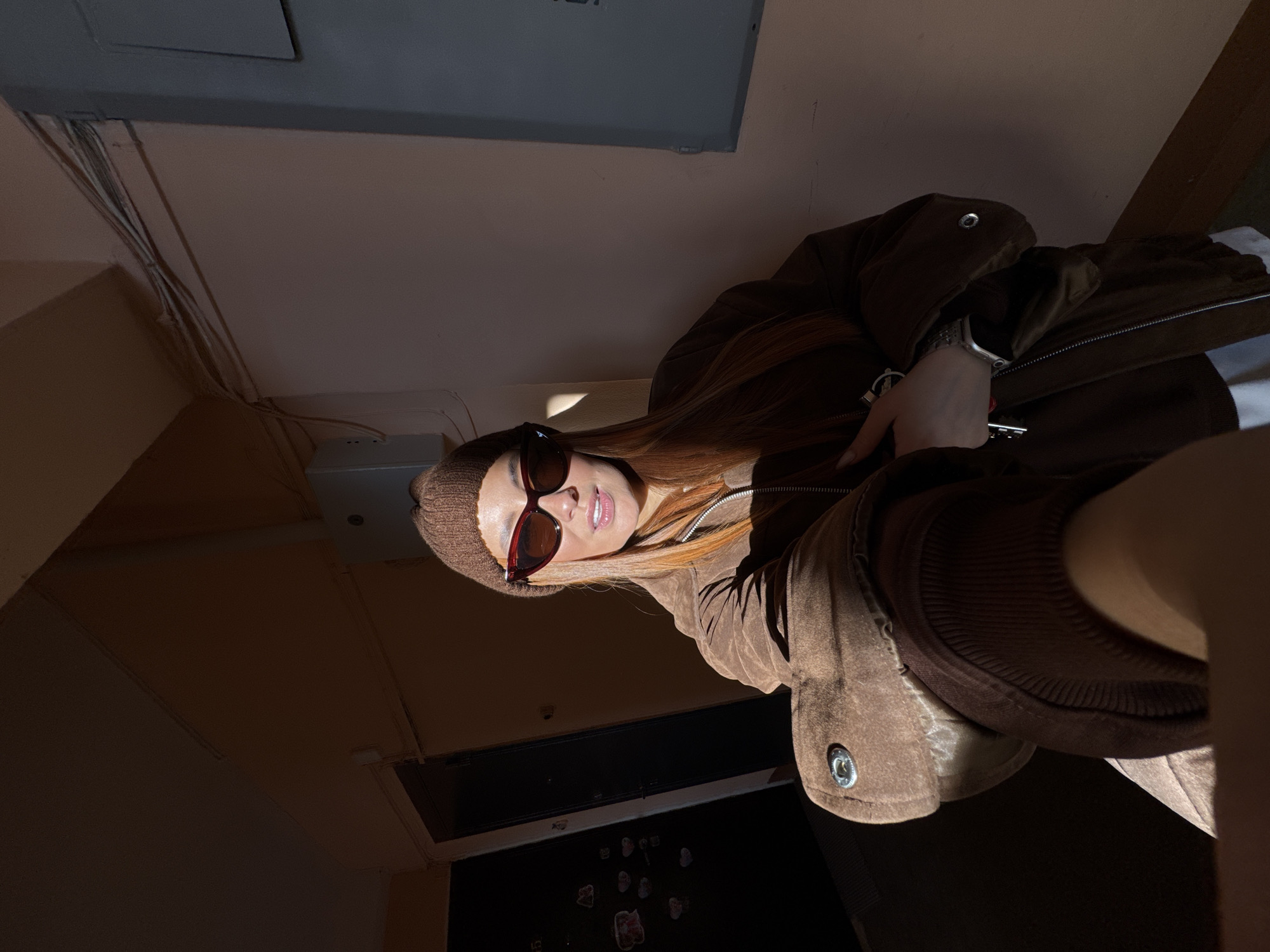 A female individual is depicted in a selfie-style photograph within a residential building hallway in Chaikovsky, Russia. She wears a brown knit beanie, dark cat-eye sunglasses with red frame accents, and a brown zippered jacket over a white garment. Her reddish-brown hair is visible beneath the beanie, and her lips are slightly parted. Her left hand holds a set of keys with a silver keychain. Intense sunlight enters from the left, illuminating the subject and casting sharp shadows. The hallway walls are a light beige. To the left of the subject, a dark apartment door is visible, decorated with several light-colored stickers, including heart shapes. To the right, a light grey rectangular utility panel is mounted on the wall, with white electrical wiring running horizontally above it.