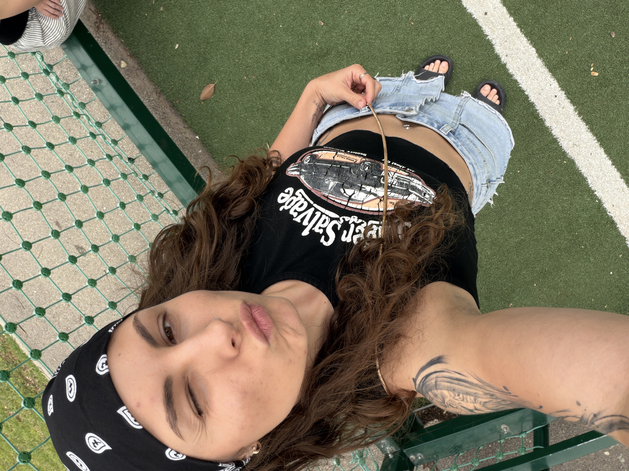 An overhead, close-up selfie captures a person lying down on a green artificial turf surface, looking upwards with a playful expression. The individual, appearing to be a young woman with long, curly brown hair, has one eye winking or squinting and her lips in a slight pout. She wears a black bandana with white paisley patterns on her head, a nose ring, and an earring.
Her attire includes a black crop top featuring a distressed graphic of a classic car or truck and the reversed text "er Salvage" (likely "Silver Salvage"). A small, reversed word "Est." is also visible. Below the crop top, her midriff is visible, showing a belly button piercing. She is wearing light blue ripped denim shorts and black slide sandals. One hand holds a thin, gold-colored necklace or chain. Tattoos are visible on her left arm and potentially her side.
The background consists of the artificial green turf with a prominent white line running across it, typical of a sports field. A concrete border and a green metal fence or netting are visible on the left side of the frame, indicating an enclosed recreational area. A dry brown leaf rests on the green turf near the fence. The lighting suggests daytime.
This scene is set in Kiryat Bialik, Occupied Palestine.