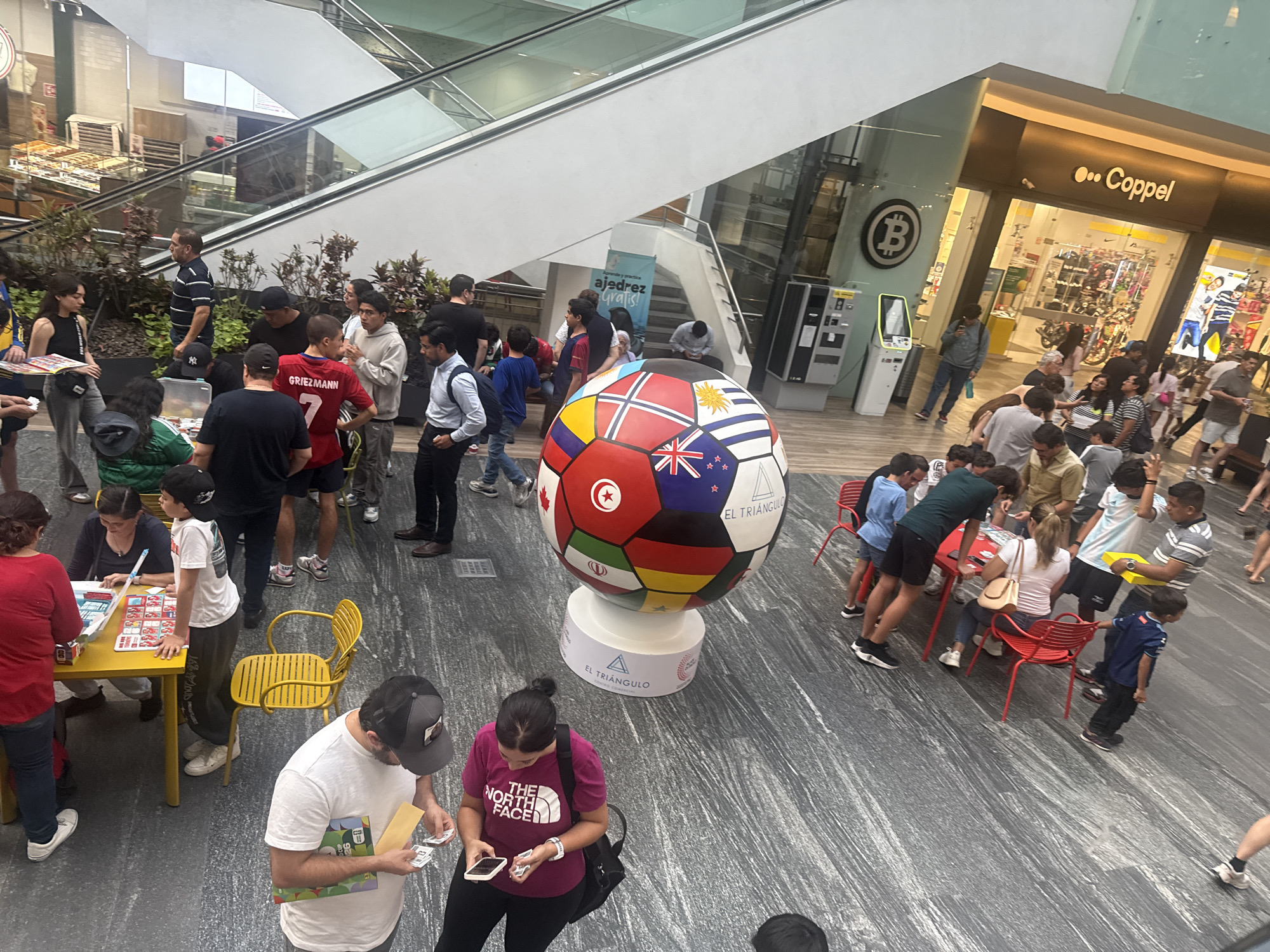The image shows a busy scene in what appears to be a mall, presumably in Puebla, Mexico, judging by the "EL TRIANGULO" logo. Many people are present, grouped around various activities. One group is gathered around a yellow table, possibly for games or crafts, another group is clustered around a soccer ball decorated with different national flags, displayed on a cylindrical stand bearing the same "EL TRIANGULO" logo. Some people are interacting with each other, looking at phones or items in their hands. Shops like "Coppel" are visible in the background. An escalator is nearby. Some individuals are sitting on red chairs at a small table. People are dressed in casual attire.