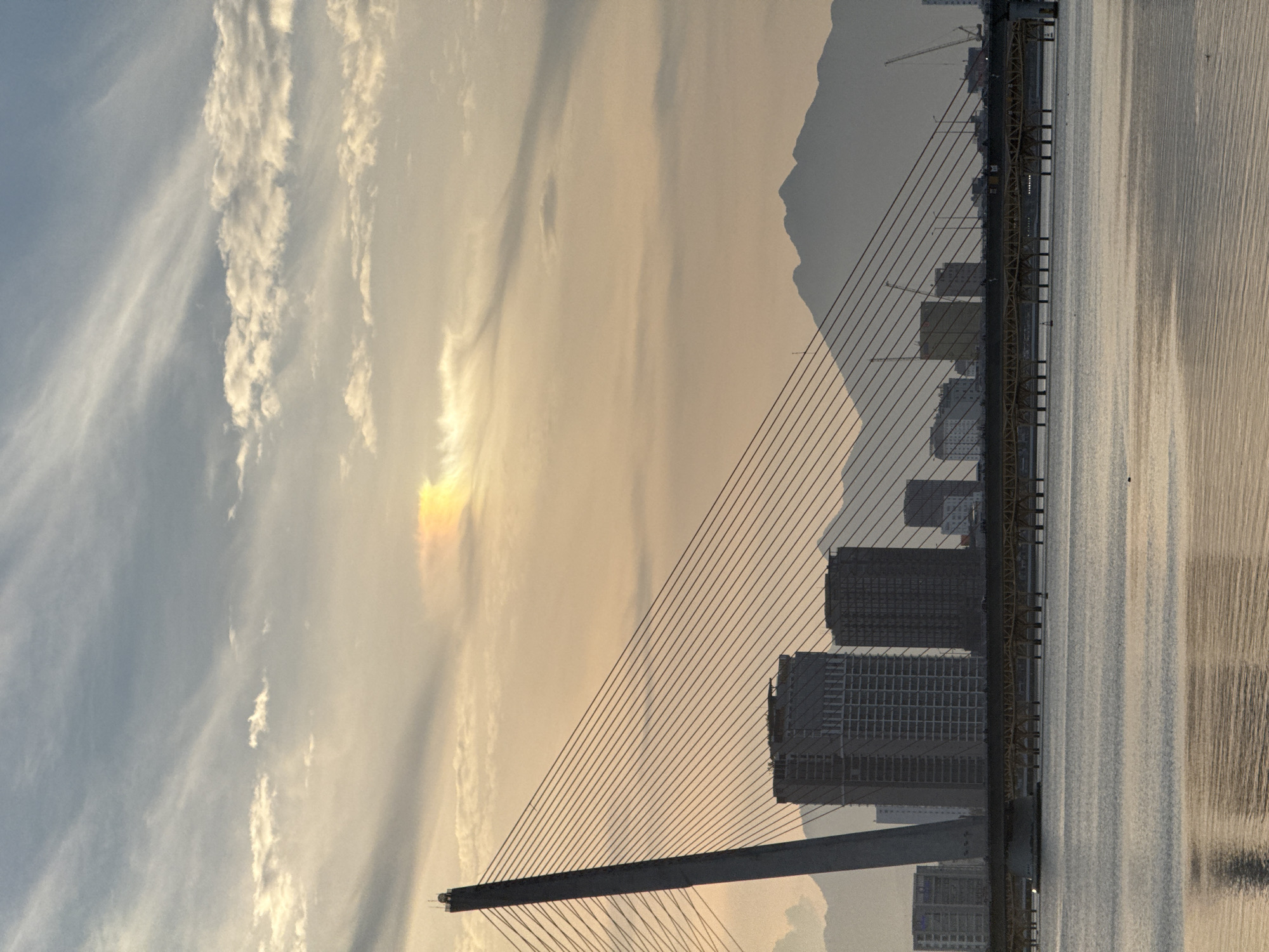 This is a photograph of the Dragon Bridge in Da Nang, Vietnam, at sunset. The bridge, a prominent landmark, is a cable-stayed bridge spanning the Han River. The image captures the bridge's distinctive architecture, with its single pylon and numerous cables extending to the roadway. In the background, the city skyline is visible, with several modern buildings and construction cranes dotted amongst them. The mountains of Da Nang form a silhouetted backdrop against the colorful sky. The water of the Han River reflects the warm hues of the sunset, creating a serene and picturesque scene. The sky is filled with wispy clouds, illuminated by the setting sun, which casts a golden glow over the entire landscape. A faint rainbow-like atmospheric optical phenomenon can be observed in the upper part of the sky.