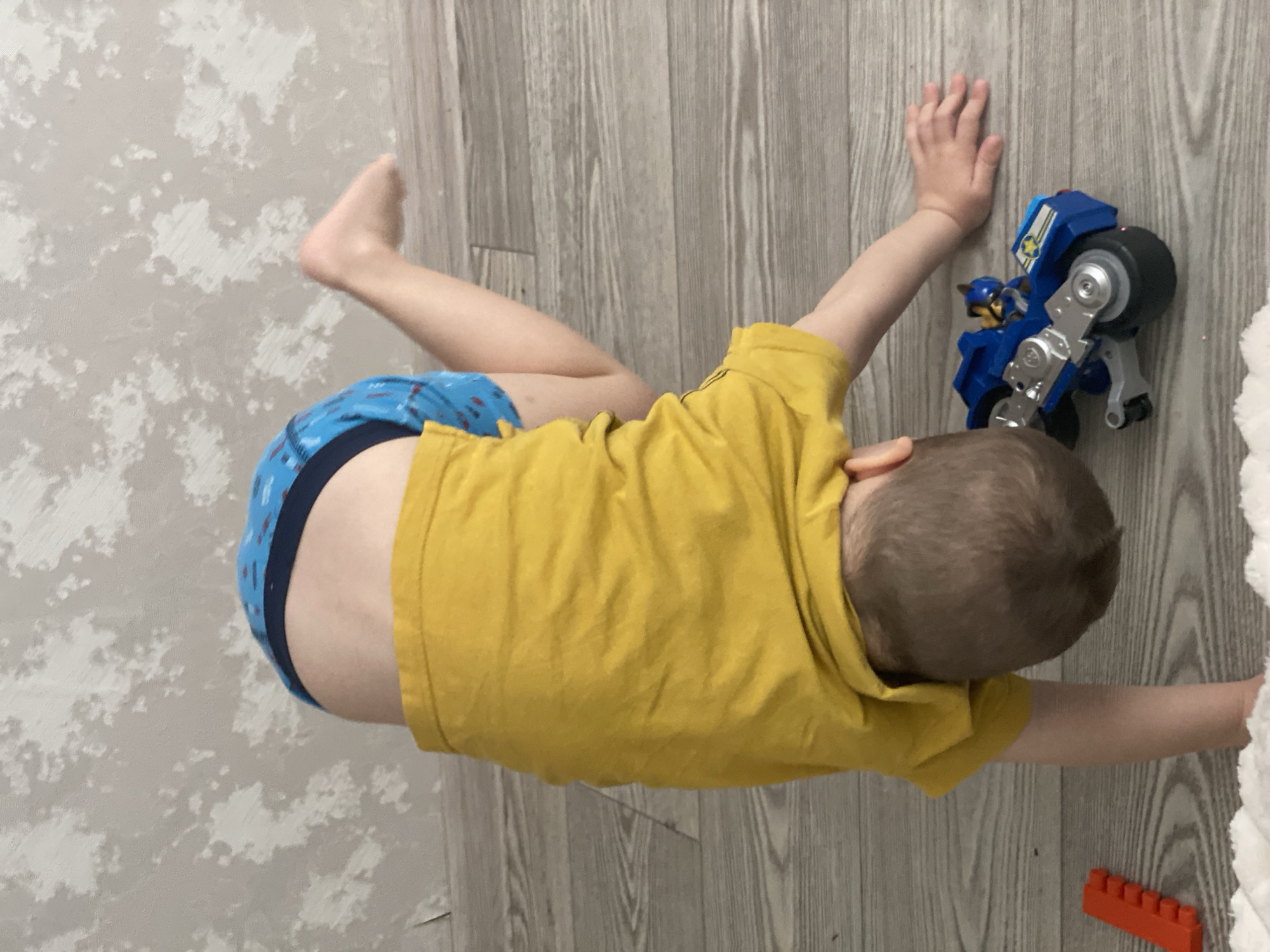 A young child is lying on a wooden floor in an interior setting, wearing blue patterned underwear and a yellow t-shirt. The child is positioned near a wall with a patterned design. A blue toy motorcycle is adjacent to the child's right hand. An orange toy brick is near the child's outstretched arm. The location is unknown and not discernible from the image.