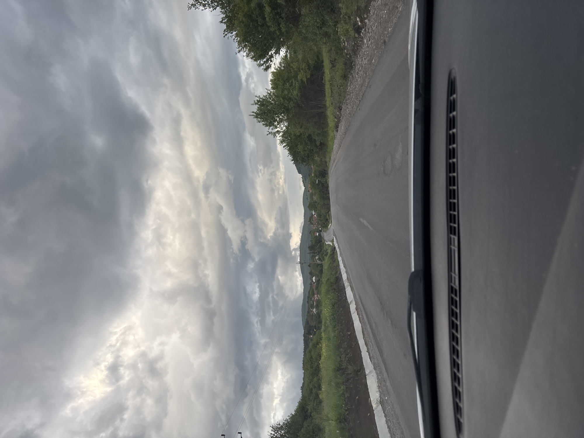 The image depicts a view from inside a vehicle, looking out onto an asphalt road in Baylovo, Bulgaria. The road stretches into the distance, flanked by vegetation on both sides, including trees and grassy areas. In the background, a village or town is visible near a hillside. The sky is overcast with gray and white clouds. The vehicle's interior, including a vent and windshield wiper, is visible on the right side of the image.