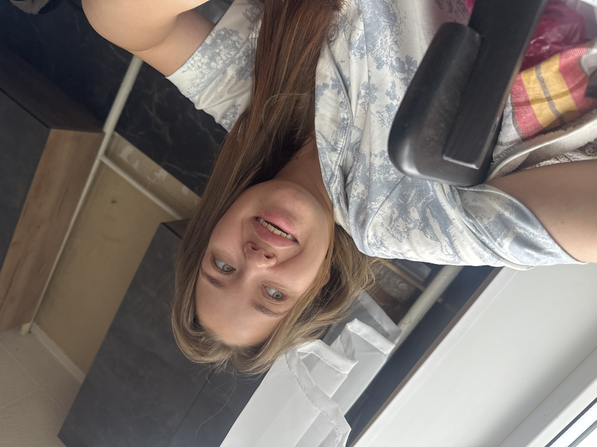 A young woman with long, brown hair and green eyes is captured in a selfie-style photo. She is wearing a light blue and white patterned pajama top. Her mouth is open in a smile, revealing her teeth. Her head is tilted to the side, and she is looking away from the camera. In the background, there are dark gray cabinets, a wooden surface, and a white curtain or blinds. The scene appears to be indoors, possibly a bedroom. The lighting suggests it is daytime, with natural light coming from the window. There is no discernible information about the location within the city of Chaikovsky, Russia, or any specific activity or interaction.