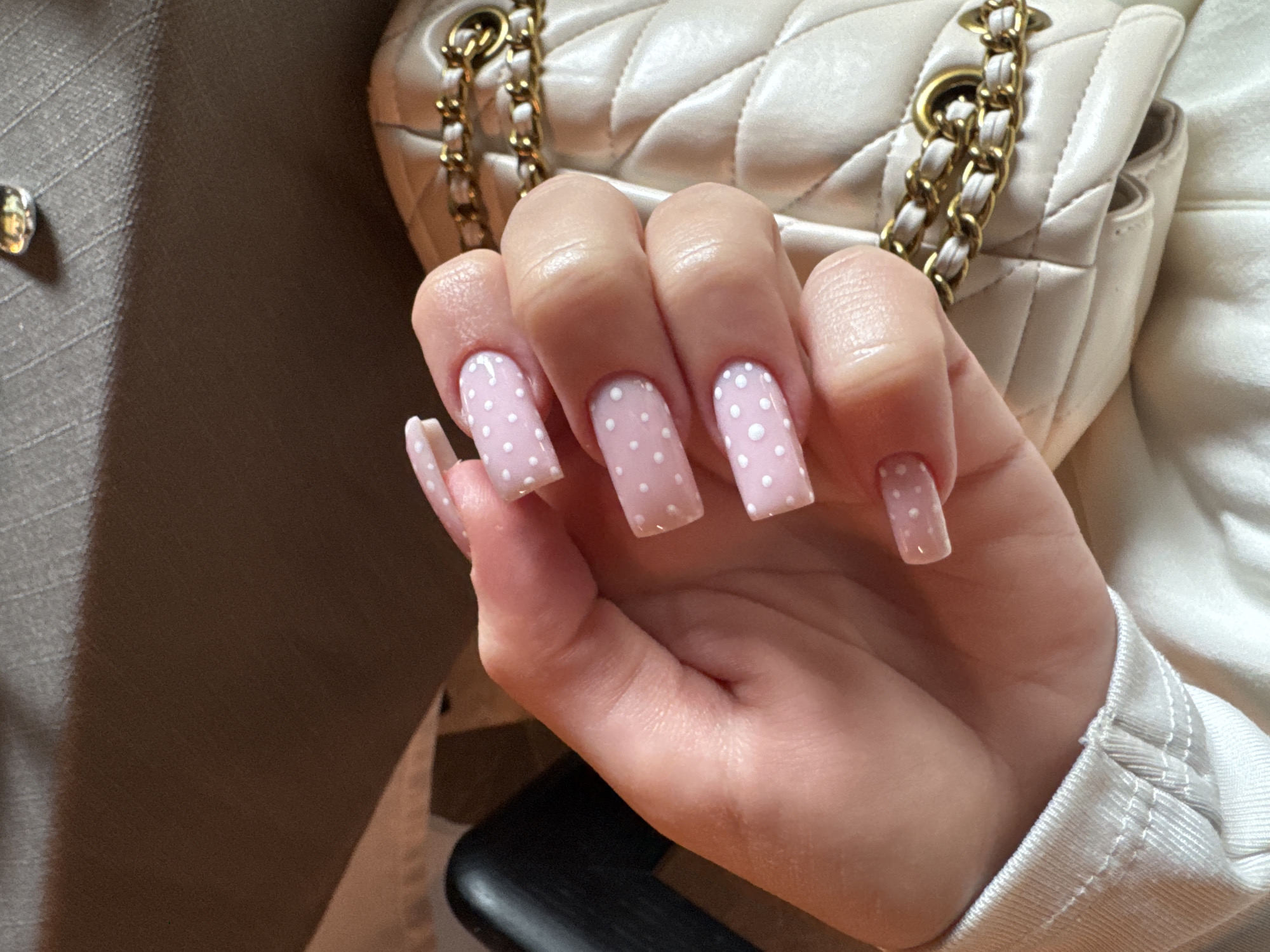 A hand with long, square-shaped nails is prominently displayed, with a light pink base color and white polka dots. The hand is holding a beige quilted purse with gold chain straps. The background includes portions of a beige garment with a visible button, and an indistinct dark object. The scene is an indoor setting. The location is not discernible from the image.