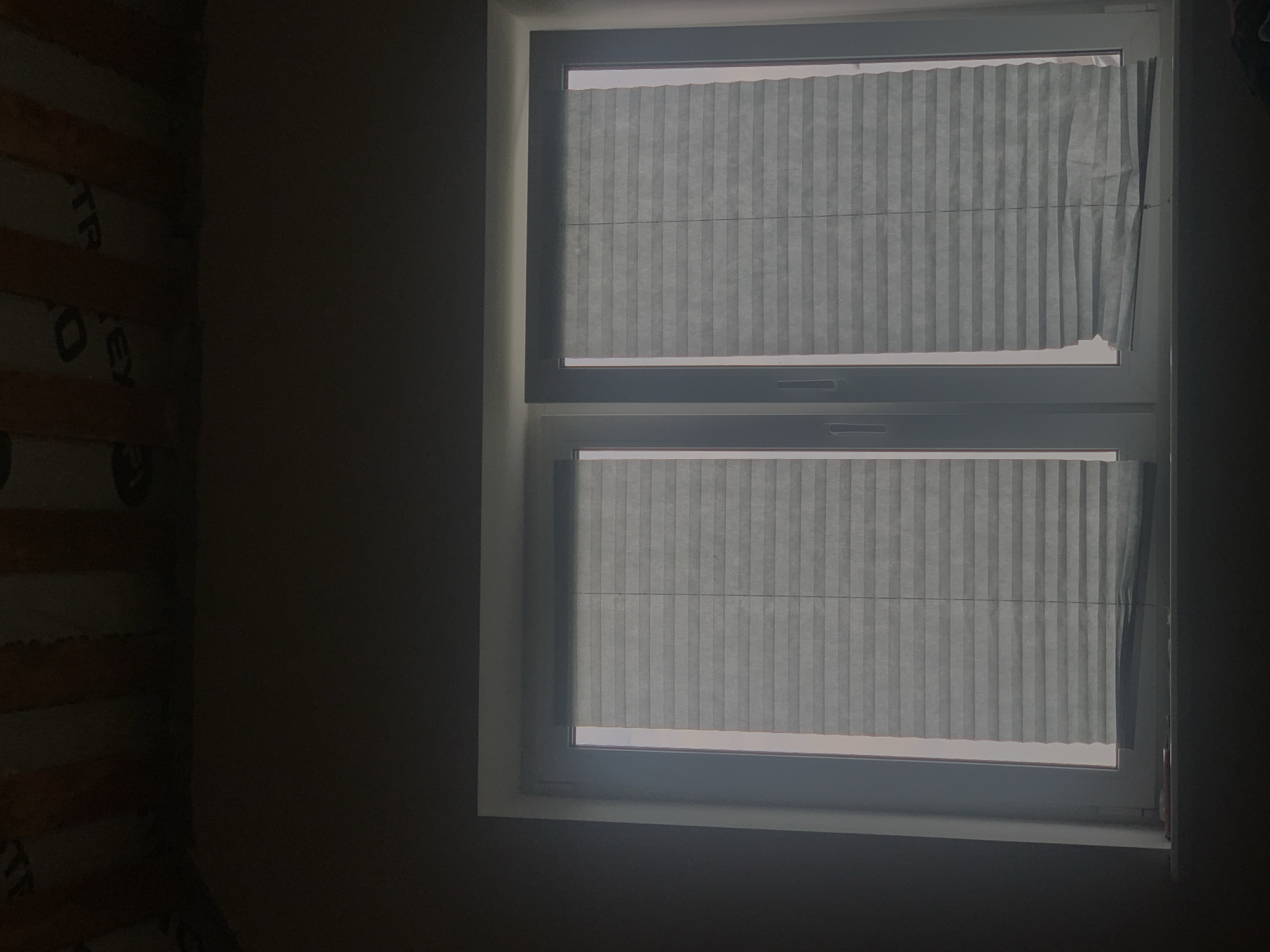 The image shows a close-up of a window with pleated blinds, seen from inside a dimly lit room. The window is divided into two panes by a vertical mullion, and each pane is covered by a light grey pleated blind. The blinds are semi-transparent, allowing some light to filter through from the outside. The right blind appears slightly uneven at the bottom, with some pleats creased or out of place. The window frame is white. Above the window, the ceiling is visible, showing wooden beams and what appears to be insulation material with some printed text, possibly from a construction product. The overall lighting is low, with the brightest area being the light filtering through the window. The scene does not contain any people or specific identifiable landmarks that would allow for localization in Minsk, Belarus.