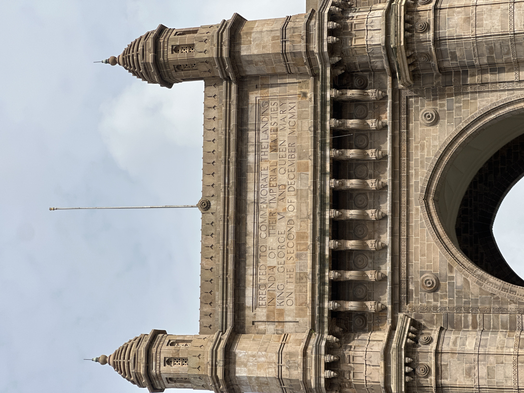 The image shows a close-up, eye-level, exterior shot of the Gateway of India in Mumbai, India. The monumental archway, constructed from warm-toned stone, is the primary subject. Carved inscriptions are visible on the facade, reading "ERECTED TO COMMEMORATE THE LANDING IN INDIA OF THEIR IMPERIAL MAJESTIES KING GEORGE V AND QUEEN MARY ON THE SECOND OF DECEMBER MCMXI". The architectural details include two octagonal turrets topped with spires on either side of the main arch, decorative carvings along the upper parapet and below the inscriptions, and corbels supporting a decorative frieze. An empty flagpole stands in the center, extending upwards into a cloudy sky. The lighting suggests it is daytime. No people are visible in the frame.