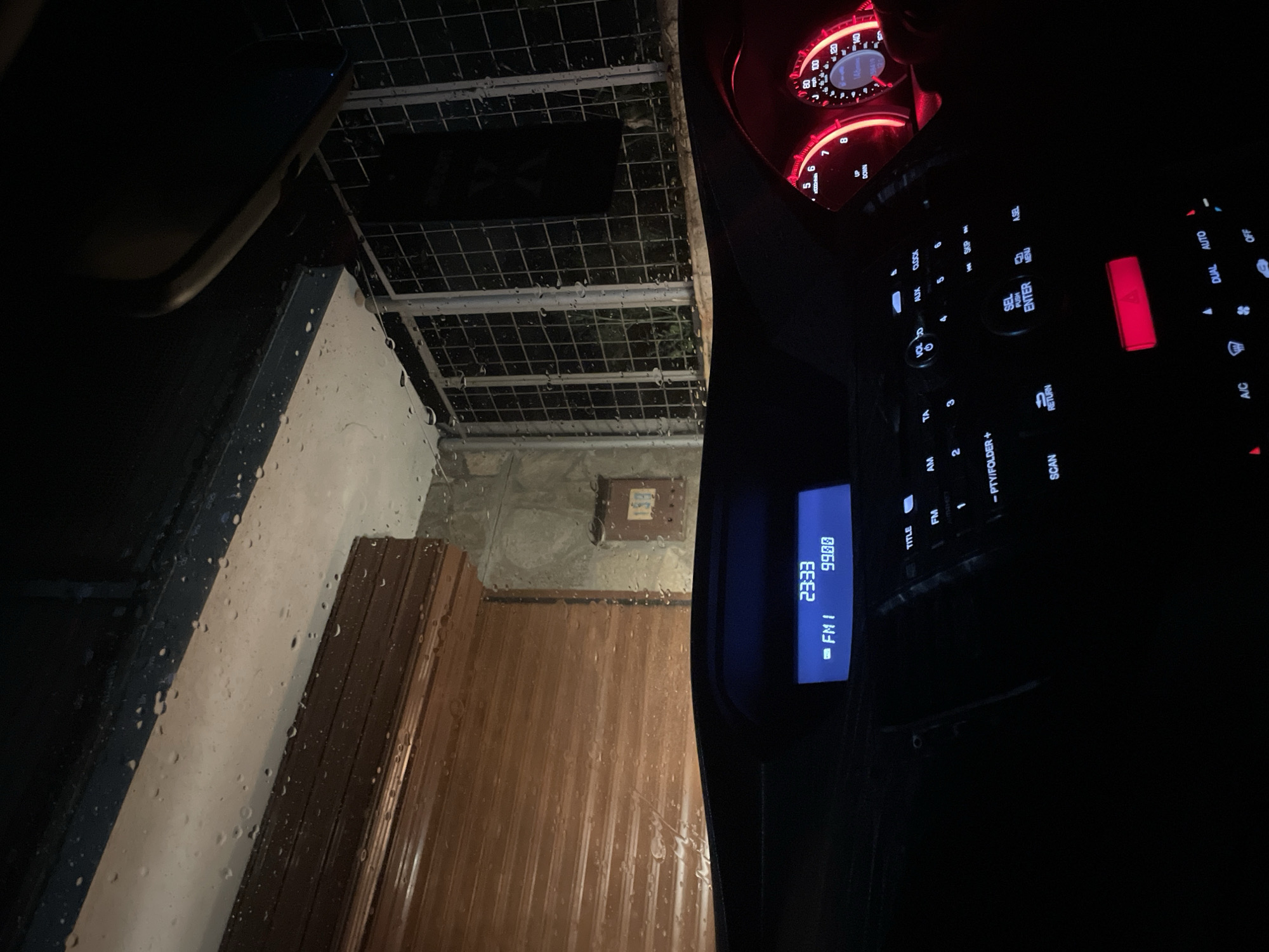 The image depicts the view from inside a vehicle, likely a car, parked near a building in Zlatograd, Bulgaria. The windshield is covered in raindrops, obscuring the view of a building with a wooden door and a concrete wall. A metal grate structure is visible above. The vehicle's dashboard is visible, including a speedometer, radio display showing "FM I 23:33 99.00", and various control buttons. The interior of the car is dimly lit, with red illumination from the dashboard. The scene is captured at night.