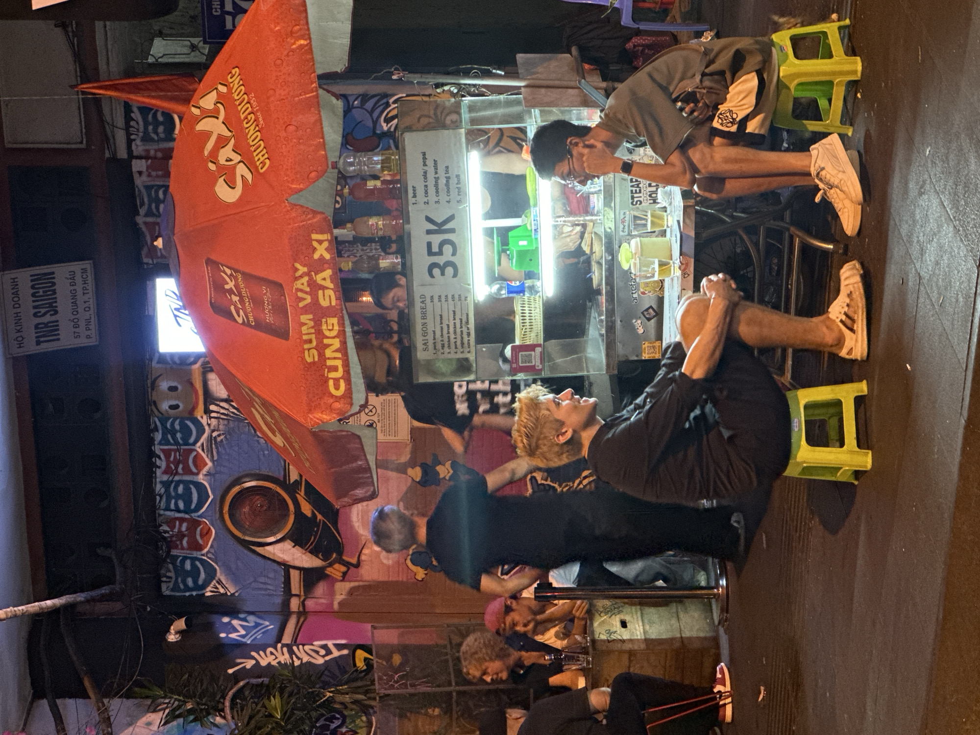 **Scene Description:**
**Location:** A street scene in the city of Thủ Đức, Vietnam, at night. The environment is urban with graffiti and street art decorating the walls.
**People:**
* A young man with blonde hair is sitting on a green stool, interacting with others.
* Another young man with dark hair is sitting at a stall.
* Other individuals can be seen in the background, sitting and engaging with the street food stall.
**Objects:**
* A street food stall is present with a glass display.
* The stall sells a variety of items, including Saigon bread, beer, coca cola, cooling water, cooling tea, and red bull.
* A large orange umbrella with the "Saxi Chuong Duong" brand covers the stall.
* Green plastic stools are used for seating.
* Graffiti and murals cover the walls in the background.
**Actions & Interactions:**
* People are seated at the stall.
* Individuals appear to be ordering or consuming food and drinks.
* There is interaction among the individuals seated at the stall.
* The stall is actively serving customers.
**Details:**
* The price "35K" is prominently displayed on the stall.
* The name "TNR Saigon" is visible on a sign.
* Graffiti is visible on the walls.
* The street is paved and dark.