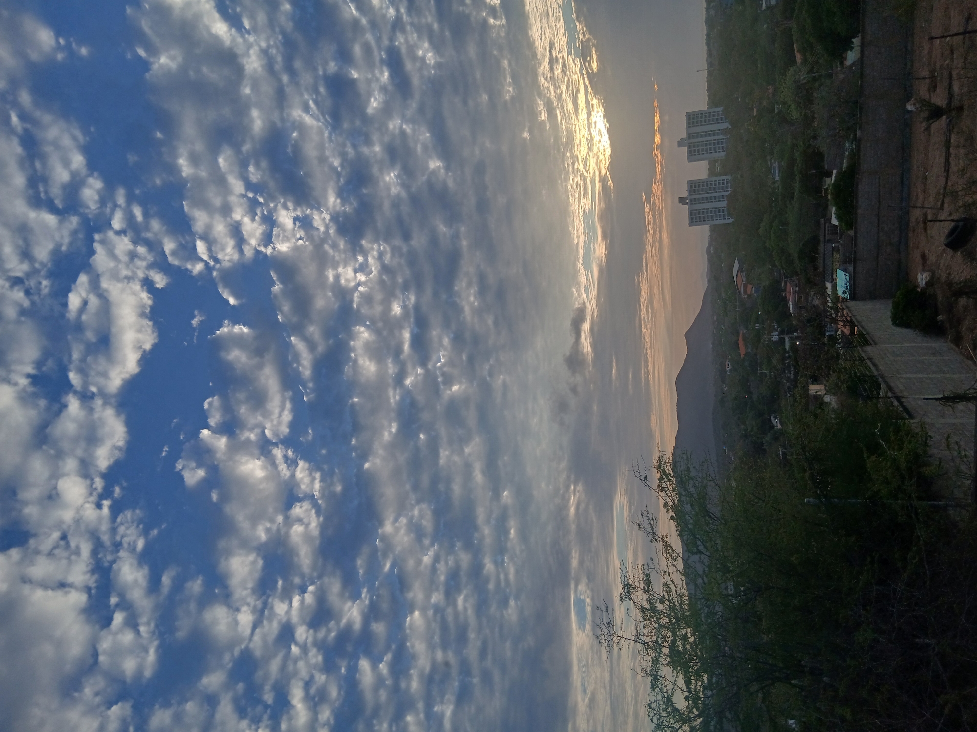 The image captures a partially obstructed view of a sunset over Juan Griego, Venezuela. The foreground is dominated by a sky filled with small, puffy clouds against a blue backdrop. Lower in the frame, trees and urban structures occupy the midground. Three tall buildings are visible on the horizon, partially obscured by the setting sun. Beyond the city is the arid landscape of arid mountains, with a peak visible in the center. A concrete wall and sparse vegetation are in the immediate foreground on the right side of the image. Overall, the scene presents a moment of transition as day turns to dusk in a coastal Venezuelan town.