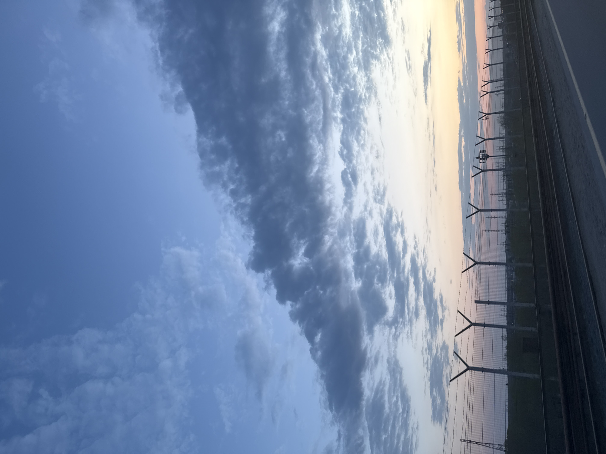 The image shows a sky with varying cloud formations, ranging from thin, wispy clouds to a thicker, more defined cloud bank. There is a subtle gradient of color in the upper right corner of the frame, suggesting the presence of the sun, either at sunrise or sunset. In the foreground, a fence is visible, constructed with vertical posts and wire mesh. Beyond the fence, there is a suggestion of industrial or infrastructure elements, possibly related to a train line. Without further context, it is difficult to definitively identify the location as Chaikovsky, Russia.