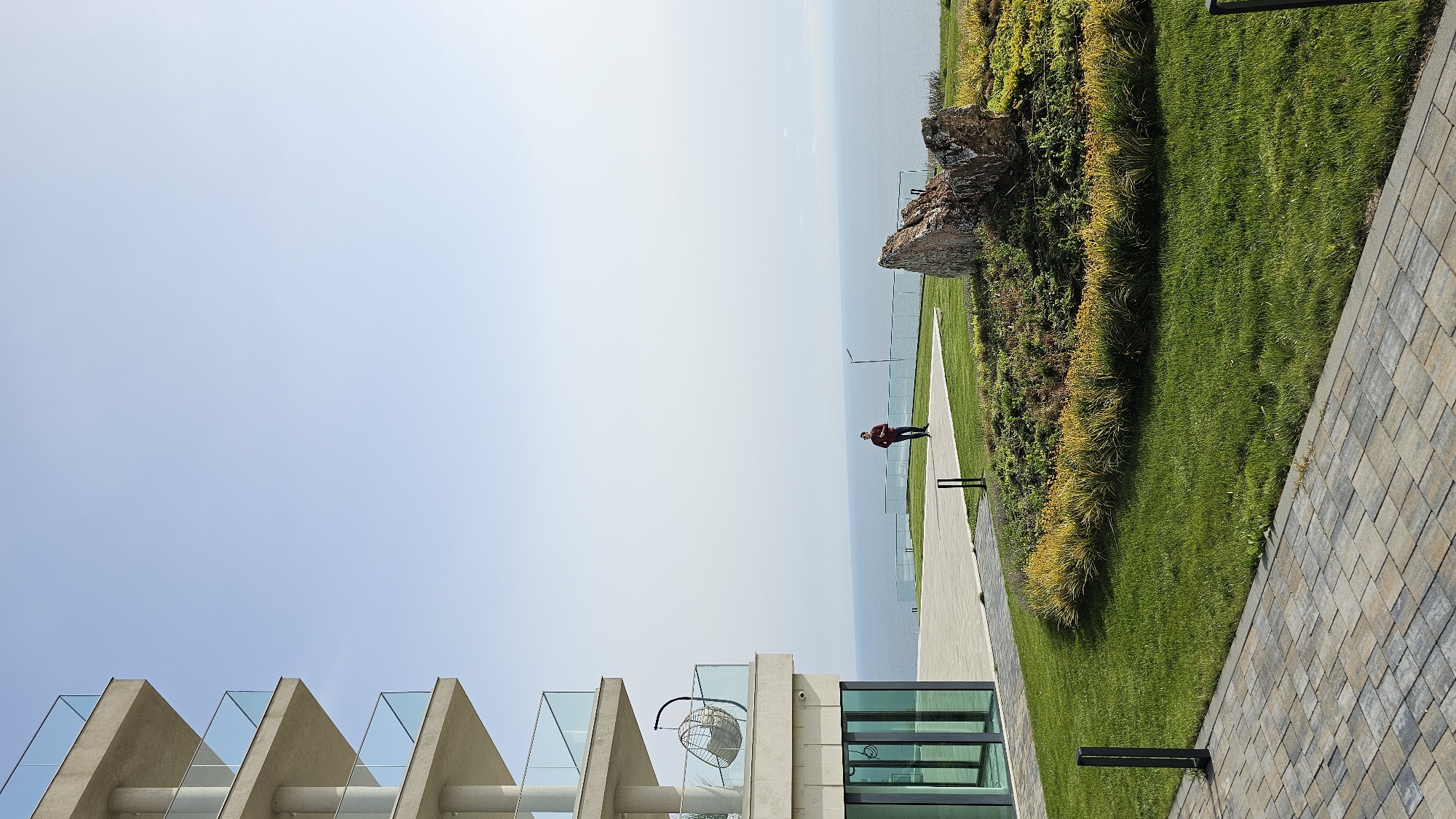 The image presents a landscape overlooking the Black Sea in Kavarna, Bulgaria. A man in a red jacket stands along a concrete walkway bordered by a low glass barrier. Beyond the barrier is the sea and sky. To the right of the walkway is a green hillside with patches of foliage. The image also features a modern building with concrete structures and glass panels in the foreground. A hanging chair is suspended from the building. A paved area made of interlocking stones is on the right of the building.