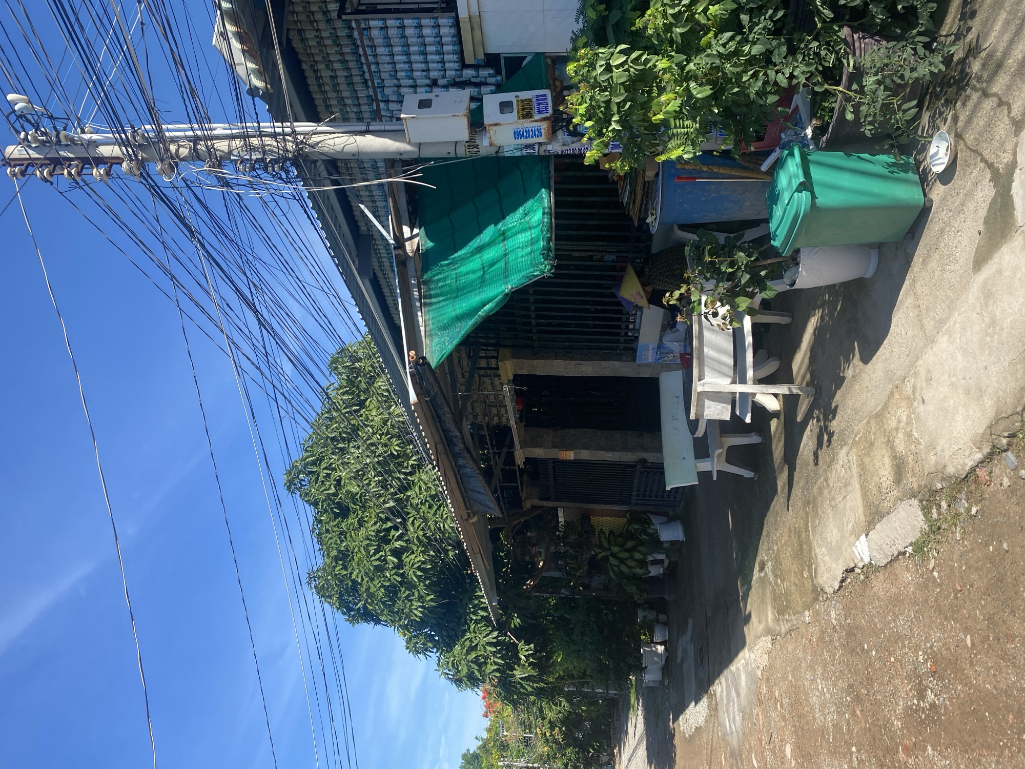 The image captures a street scene in Mũi Né, Vietnam, under a clear blue sky. The foreground shows a dirt and concrete path leading past a building. To the right, a white bench and table are situated on the concrete. The building's facade is adorned with rows of white cans, stacked to the roofline, and a corrugated metal roof extends over the front area, partially covered by a green mesh tarp. A utility pole with numerous tangled wires stands to the right of the building, and a green trash bin is visible in front of the structure, beside several potted plants. The background features lush green trees and foliage. A person wearing a conical hat is partially visible sitting near the building's entrance. The overall impression is of a residential or commercial street with dense vegetation and overhead wiring.