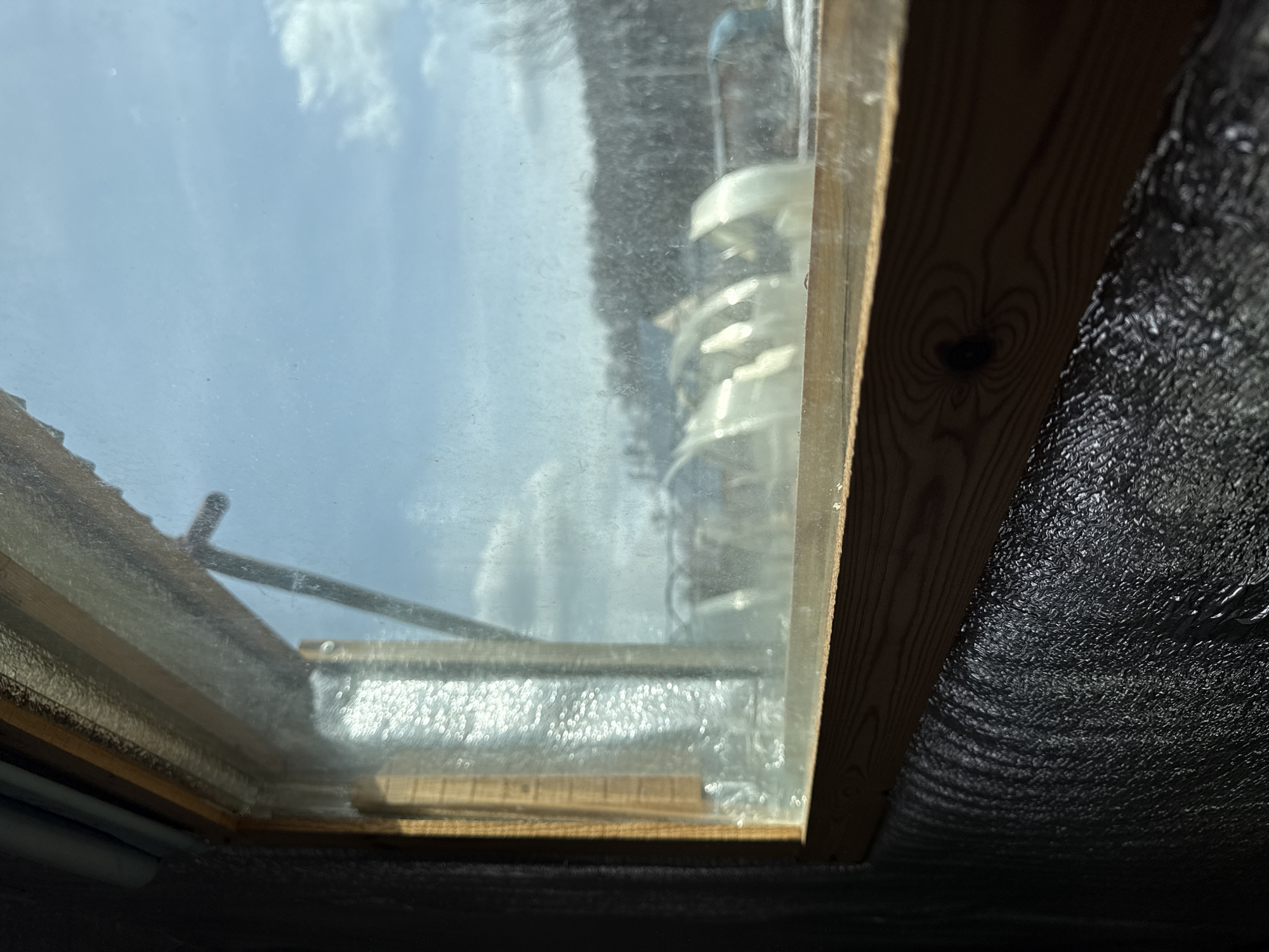 The image shows a skylight window with a wooden frame. Outside the window, the sky is visible with some clouds. The glass appears to be dirty or wet, possibly with condensation. To the right of the window frame, there is a dark, textured surface, possibly some kind of insulation material. The setting appears to be indoors, looking up at the window. There's no visible indication of location in the city of Chaikovsky, Russia.