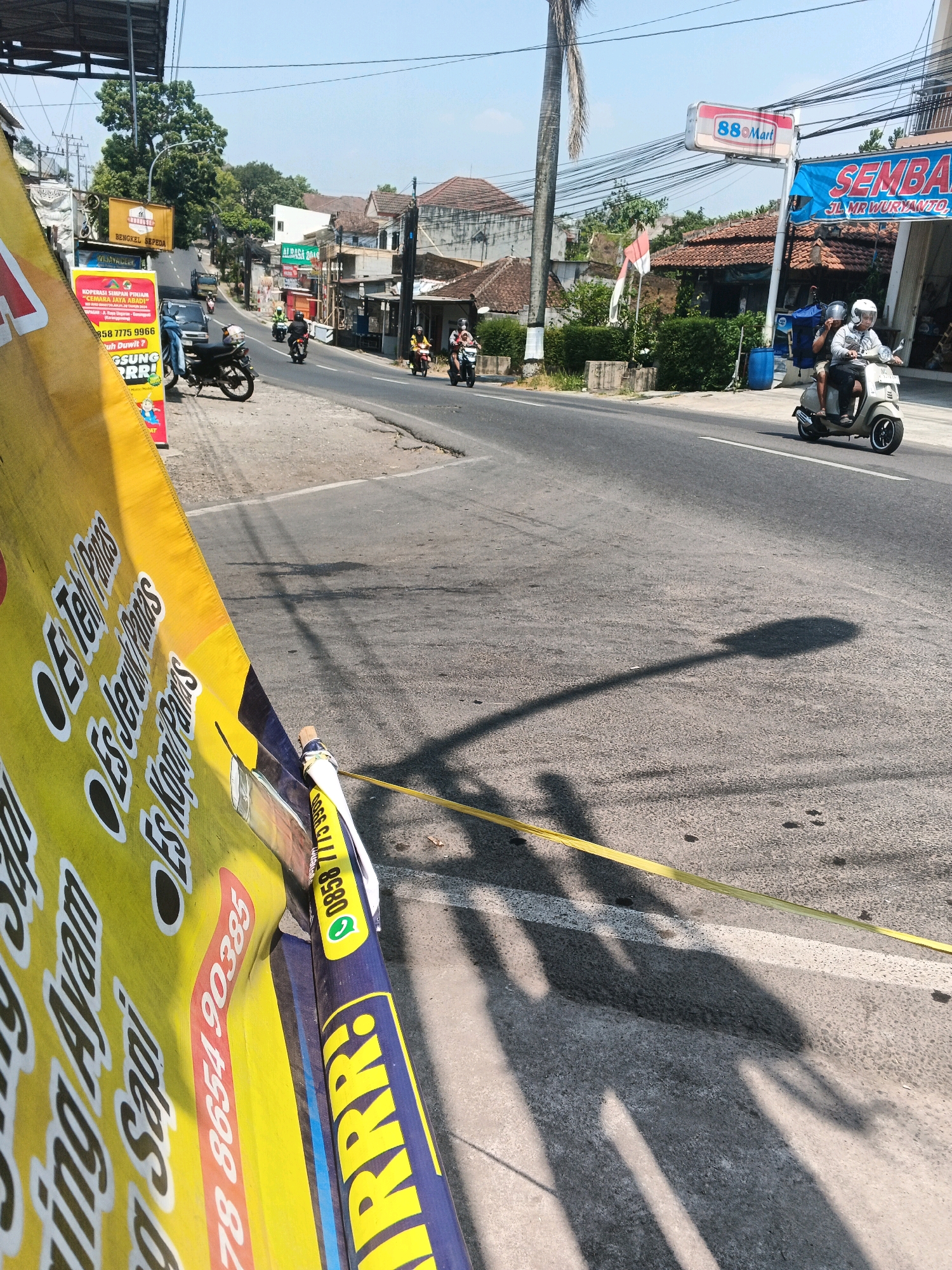 This is a street-level, medium-angle, eye-level shot taken on a sunny day, likely in the city of Semarang, Indonesia. The scene captures a busy road with traffic and pedestrian activity. In the foreground, a yellow banner advertising drinks like "Es Teh Panas," "Es Jeruk Panas," and "Es Kopi Panas" is prominently displayed, partially obscuring the view of the road. The banner also includes a phone number: "0858 7775 9966" and "0858 7775 9966," along with "78 8654 90385". On the right side of the banner, a smaller blue banner reads "RRR!".
The road is asphalt with visible tire marks and some debris. Several motorcycles are present on the road. One motorcycle, ridden by a person wearing a helmet, is in the mid-ground heading away from the camera. Another motorcycle with a rider also wearing a helmet is closer to the foreground, moving in the same direction. Further down the road, more motorcycles are visible, including one carrying two individuals.
On the right side of the road, there are buildings with signs. One sign reads "88 Mart," and another adjacent banner is blue and says "SEMBA." There's also a white sign with "JL MR WURYANTO." The architecture suggests a commercial or residential area.
In the background, buildings with tiled roofs are visible, along with trees and utility poles with numerous wires crisscrossing. The sky is clear and blue. Shadows of objects and people are cast on the road, indicating the direction of the sun. The overall impression is of a typical urban street scene in Indonesia on a bright day.
