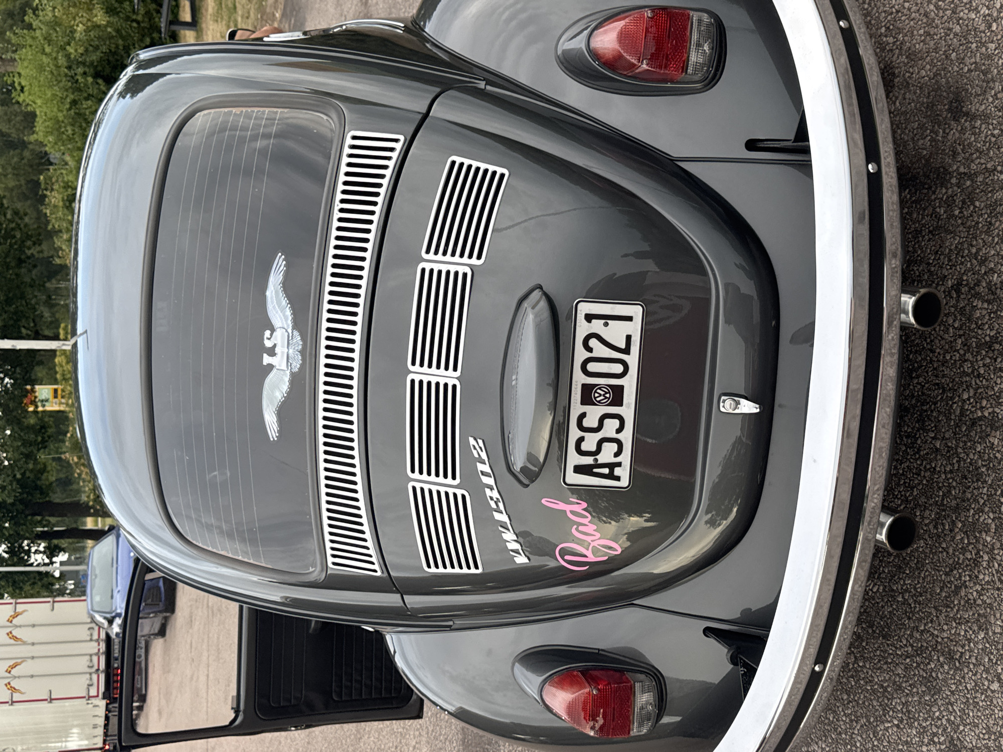A dark gray Volkswagen Beetle is parked on asphalt in Skrea, Sweden. The car's rear is facing the camera, and the driver's side door is open. The car has several stickers on its rear: a winged emblem with the letters "LS" at the top of the rear window, the text "VW 1302" below it, and a pink cursive sticker that reads "Bad" to the left of the license plate. The license plate is visible and reads "ASS 021". A trailer truck and a blue car are parked behind the Beetle. The environment appears to be an outdoor parking area with trees and vegetation in the background.