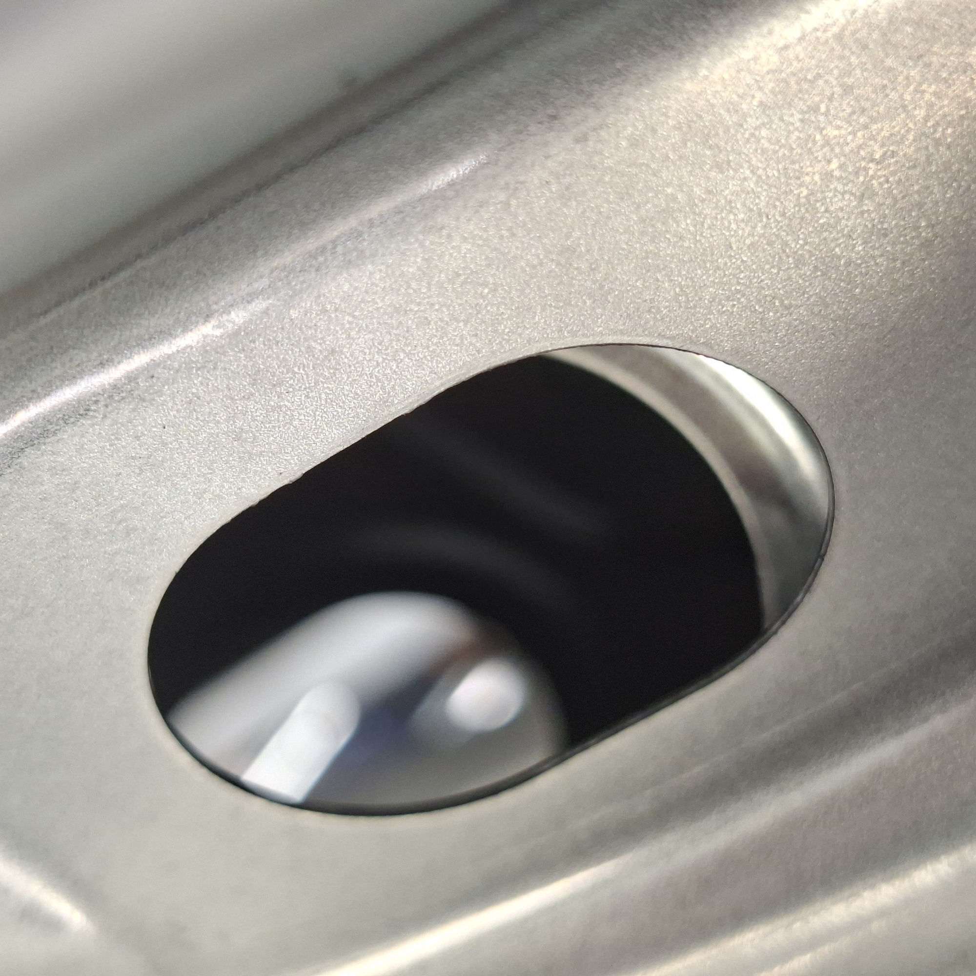 The image is a close-up, macro shot of a metal object with a textured surface and an oval-shaped opening. The metal appears to be brushed aluminum or a similar material, with visible grain and subtle variations in sheen due to the lighting. The oval opening reveals a dark, indistinct interior, possibly a cavity or a shadowed space. The foreground is dominated by the textured metal, with a strong diagonal line formed by a raised ridge or curve on the surface. The lighting creates highlights and shadows that emphasize the contours and texture of the metal. The context of Rastatt, Germany, is not visually represented in the image.
