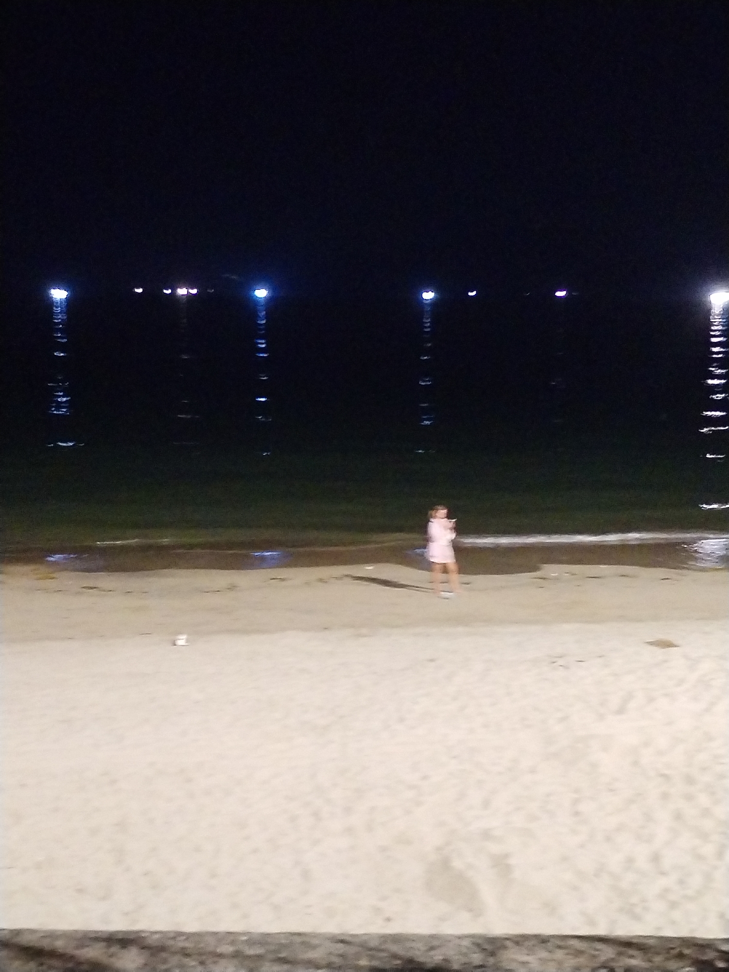 The image depicts a nighttime scene on a beach in Nha Trang Ward, Vietnam. In the foreground, there's a sandy beach. In the middle ground, a person in light-colored clothing is standing at the edge of the water. In the background, the dark water is visible with light reflections on the surface, originating from city lights on the opposite shore. The sky is dark and clear, without any visible celestial objects.