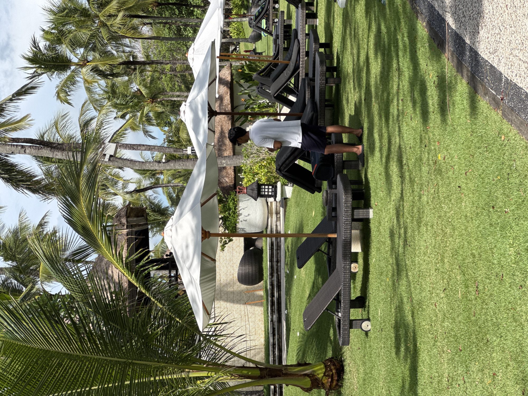 The image shows an outdoor resort area in Kuta Selatan, Indonesia. There are multiple dark-colored lounge chairs with white umbrellas on a green lawn. A person wearing a white shirt and dark shorts is standing near the chairs. Palm trees are visible in the background. A building is seen behind the lounge chairs with decorative elements. The area appears to be well-maintained.