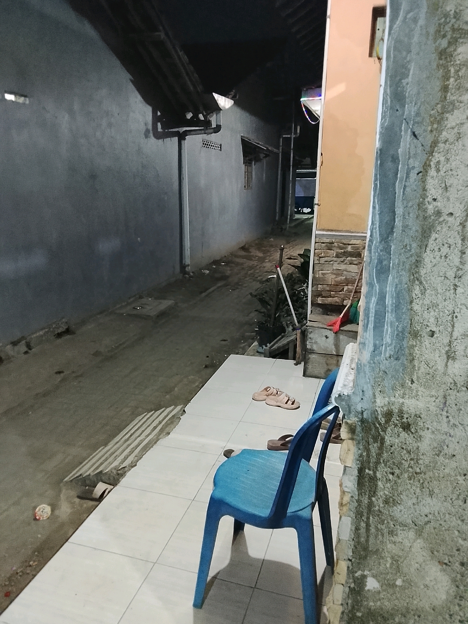 The image depicts a narrow alleyway at night in Ujungrusi, Indonesia, with a blue plastic chair in the foreground. The alley is flanked by walls on both sides. The left wall is a plain gray concrete surface, while the right features a light orange-colored building with exposed brickwork at the lower section.
On the tiled ground to the left of the chair, a pair of light brown sandals are placed side-by-side. Behind these, another pair of darker brown sandals is partially visible. To the left of the alley floor, there is a section of concrete with what appear to be metal gratings or bars, bordering a dirt path. Some debris is scattered on the ground.
In the alleyway itself, a broom with a red head and a mop with a green head are leaning against the right-side building. Further down the alley, there are some plants and what looks like a wooden structure. The overall lighting suggests it is nighttime, with artificial light sources casting shadows. The environment appears to be a residential or semi-urban area, given the presence of simple structures and the alleyway setup.