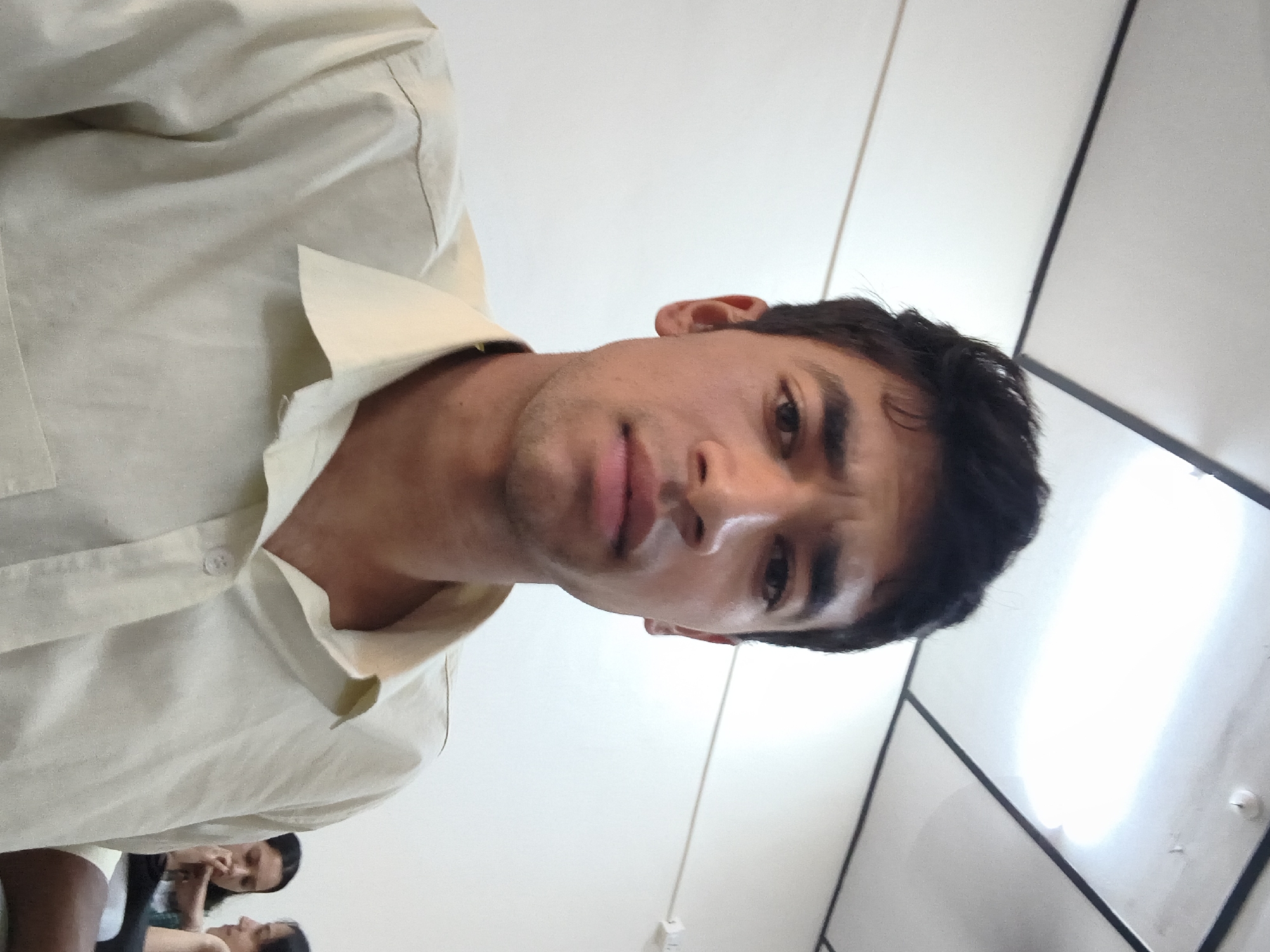 The image shows a close-up, selfie-style shot of a young man wearing a light-colored collared shirt. He has dark hair, dark eyebrows, and a serious expression. The background appears to be an indoor setting with a white wall and a ceiling with fluorescent lighting. Two other individuals, presumably women, are visible in the background, slightly out of focus, seated to the right. The man is the primary subject, filling most of the frame. The scene does not offer any visual cues to suggest a specific location within Dharamshala, India, beyond it being an interior space.