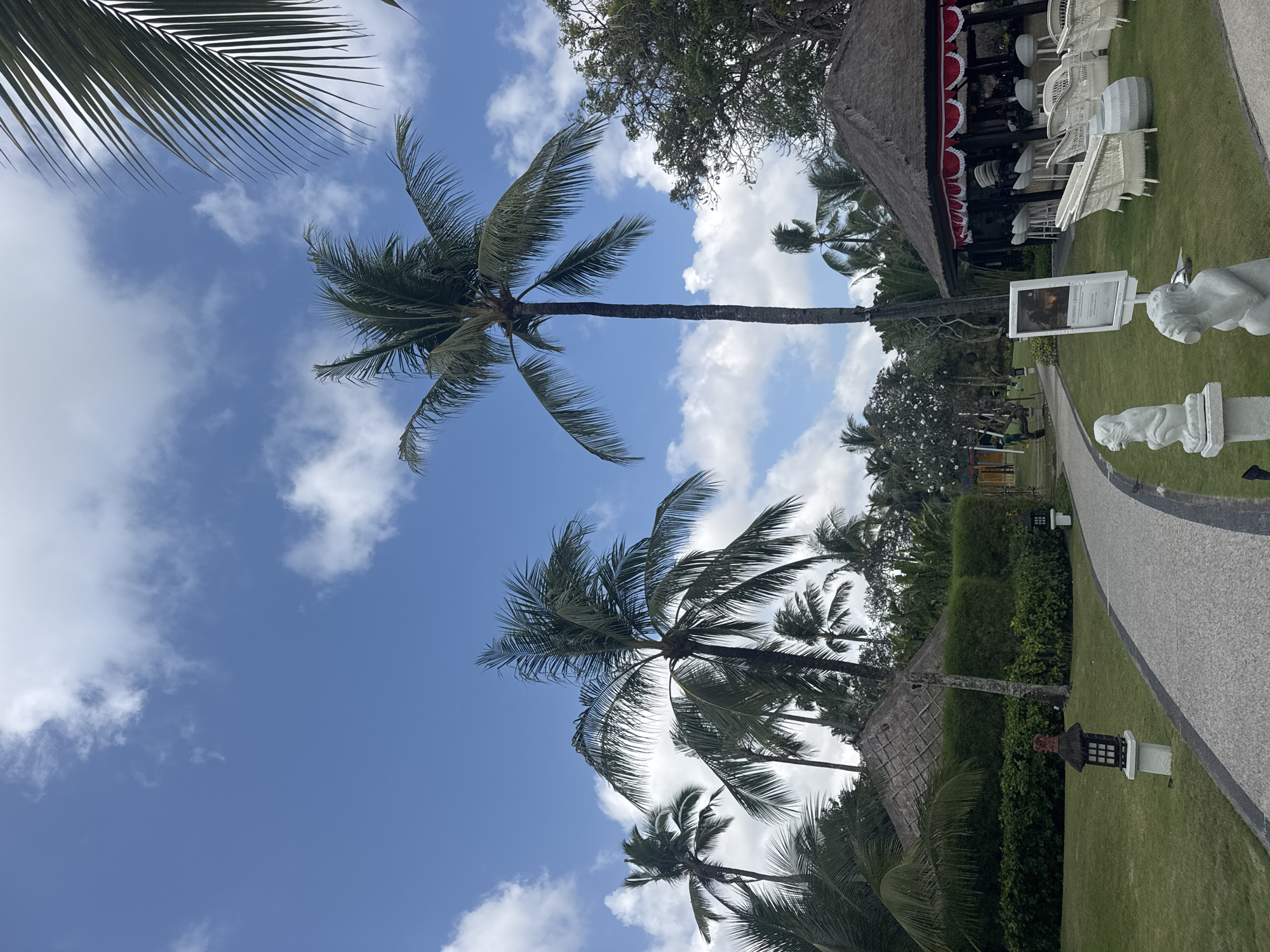 The image depicts a tropical resort scene in Kuta Selatan, Indonesia. The foreground features a paved pathway curving through a well-manicured lawn dotted with white, stylized monkey sculptures. To the left, a traditional lantern with a thatched roof sits on a stone base, beside a neatly trimmed hedge and a thatched-roof building. In the mid-ground, several palm trees with lush green fronds tower over the landscape. To the right, an open-air pavilion with a thatched roof and red and white decorative trim houses a bar area with white stools. White wicker chairs and tables are arranged on the lawn in front of the pavilion. The background is filled with more palm trees and lush vegetation under a bright blue sky with scattered white clouds. The scene suggests a serene and upscale vacation destination.