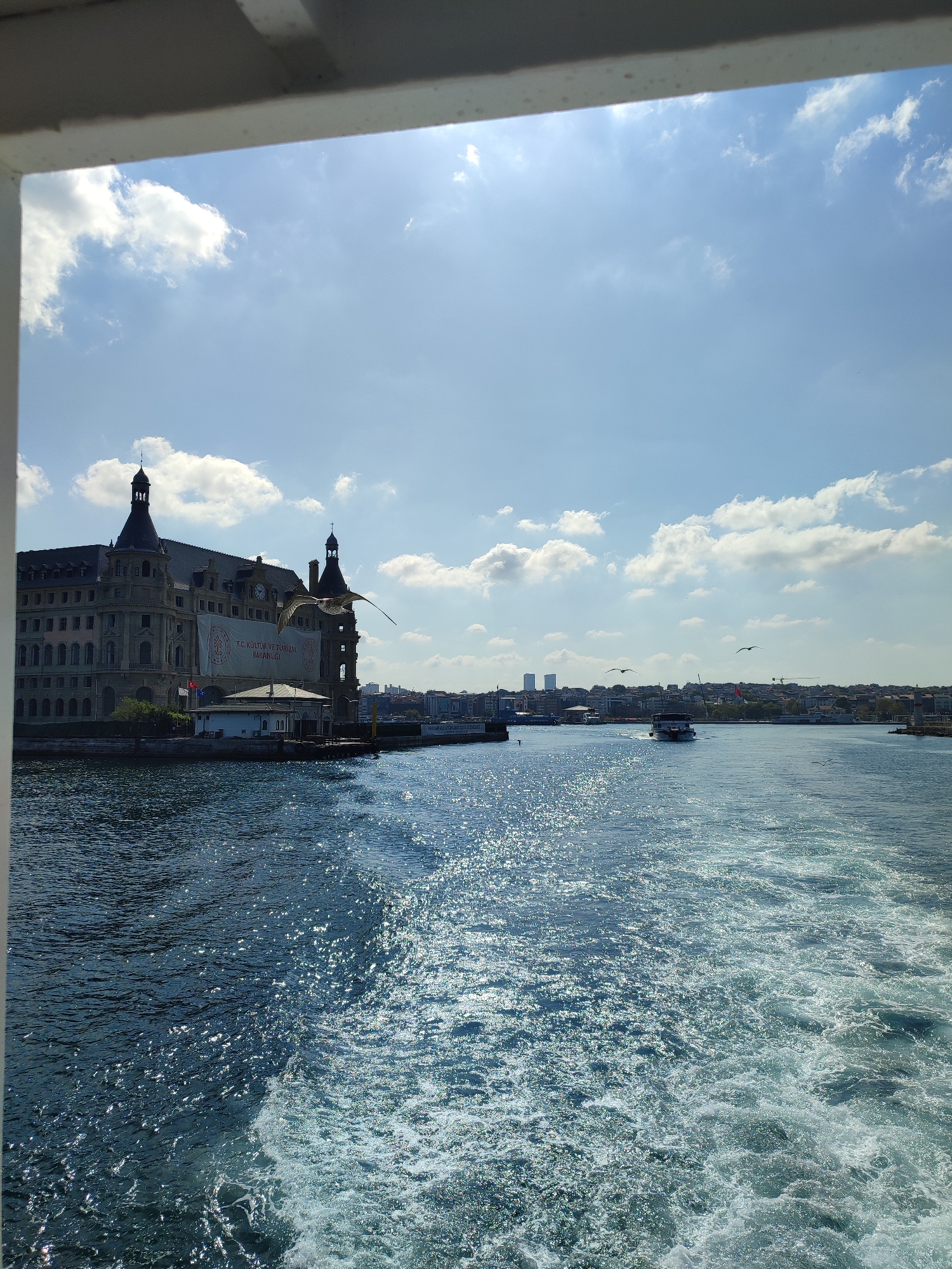 The image shows a bright, sunny day with a clear blue sky dotted with fluffy white clouds. The foreground is dominated by the deep blue water of a body of water, likely the Bosphorus Strait. A white wake trails behind the viewer's vantage point, indicating movement through the water.
On the left side of the image, a large, ornate building with distinctive domed towers and numerous windows stands prominently along the waterfront. A large banner with Turkish writing hangs on the building's facade. In front of this building is a smaller white structure with a red-tiled roof, and a pier extends into the water. Seagulls are visible in flight.
Further in the distance, along the coastline, is a city skyline with buildings of varying heights, including some taller modern structures. A ferry or boat is navigating the water in the mid-ground. The sun is shining brightly, creating a shimmering effect on the water's surface. The overall impression is a scenic view of a coastal city from the water. This building on the left is Haydarpaşa Terminal, a historic railway station located in the Kadıköy district of Istanbul, Turkey.