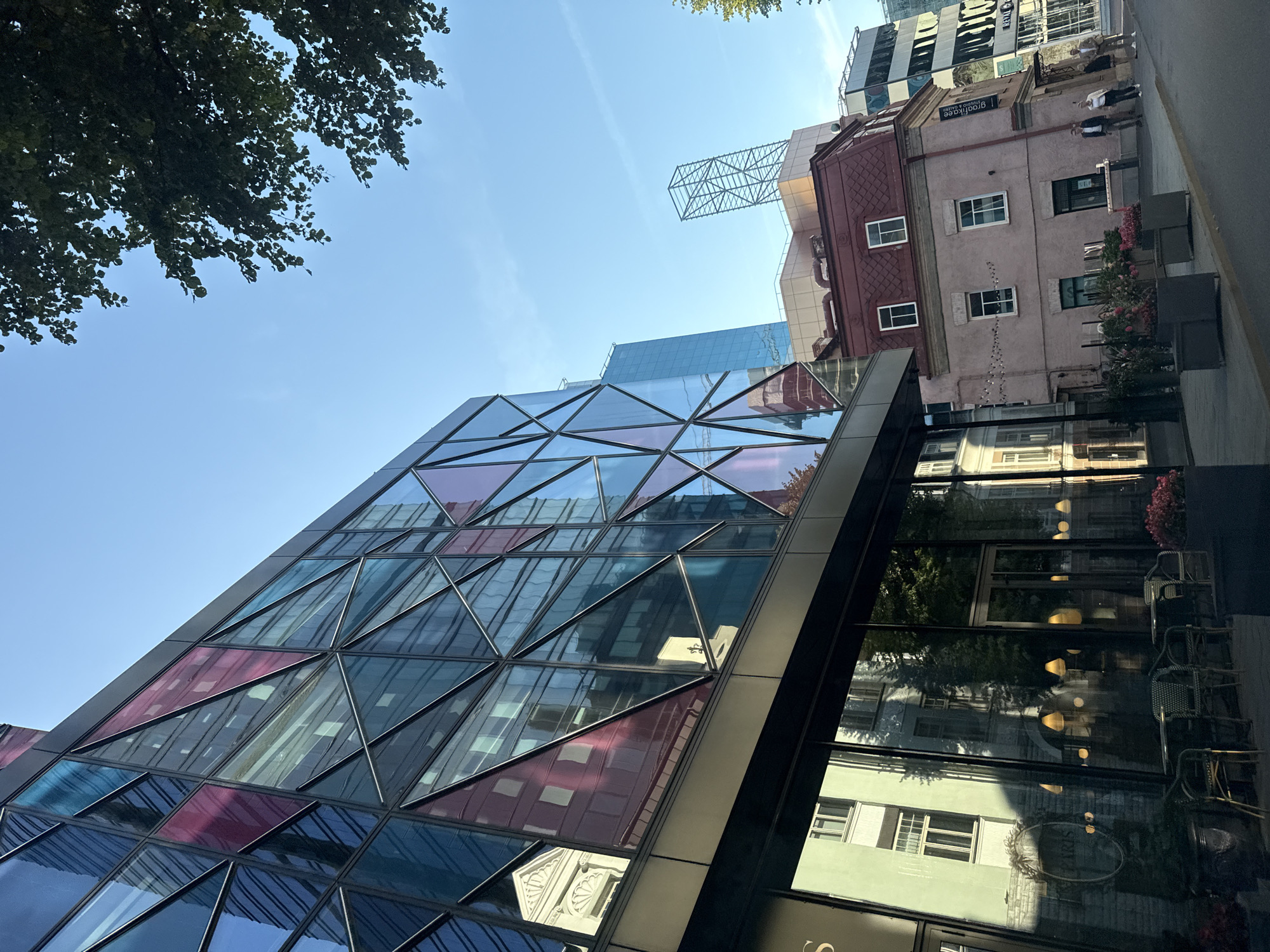 The image shows a street scene in Tallinn, Estonia. A modern building with a facade of triangular glass panels in various shades of blue and pink dominates the left side of the frame. These panels reflect the surrounding buildings and the sky. Below this modern structure, a commercial establishment with large glass windows and outdoor seating is visible. To the right of the modern building, a smaller, older building with a mansard roof and a light pink facade stands. Several people are walking on the sidewalk in front of this building. Further to the right, another modern building with a black and white striped facade is partially visible. The sky is clear and blue, with a few faint contrails. Green trees overhang the scene from the upper right corner. The overall impression is of a contemporary urban environment with a mix of old and new architecture.
