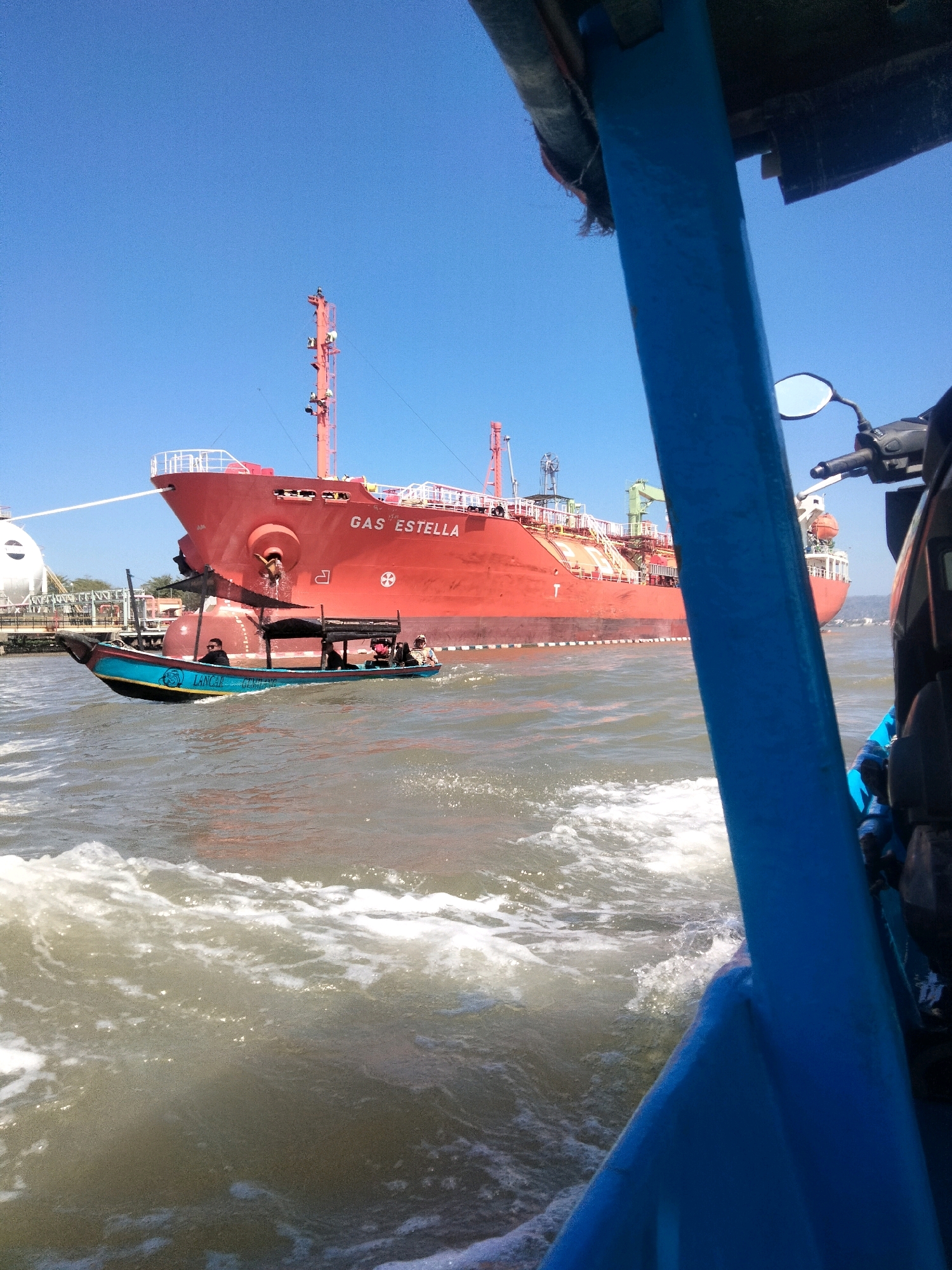In Desa Lomanis, Indonesia, a large red tanker ship, labeled "GAS ESTELLA," is anchored in a body of water. In the foreground, a smaller blue and black boat with a canopy is moving on the water, creating a wake. Several people are visible in the smaller boat, one of whom is looking towards the tanker. The sky is clear and blue. Part of a blue railing and a motorcycle are visible in the immediate foreground, suggesting the image is taken from another boat or vessel.