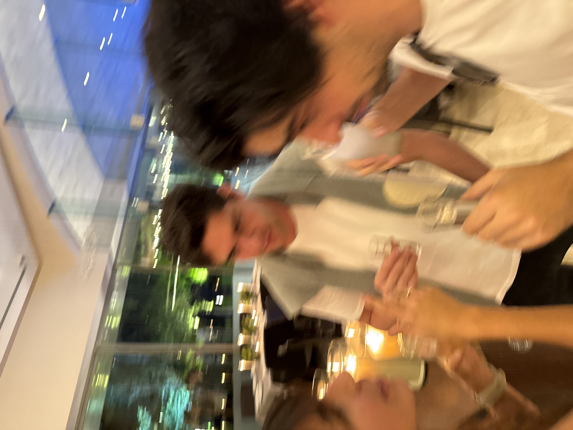The image shows a group of people at a table inside a building, likely a restaurant, with large windows overlooking an outdoor area that may be a garden or terrace. The location appears to be in Mexico City, Mexico. The people are toasting or celebrating, with several hands visible holding glasses. The table is set with candles. There are three people visible. A building and vegetation are visible outside the windows. The image is blurry.