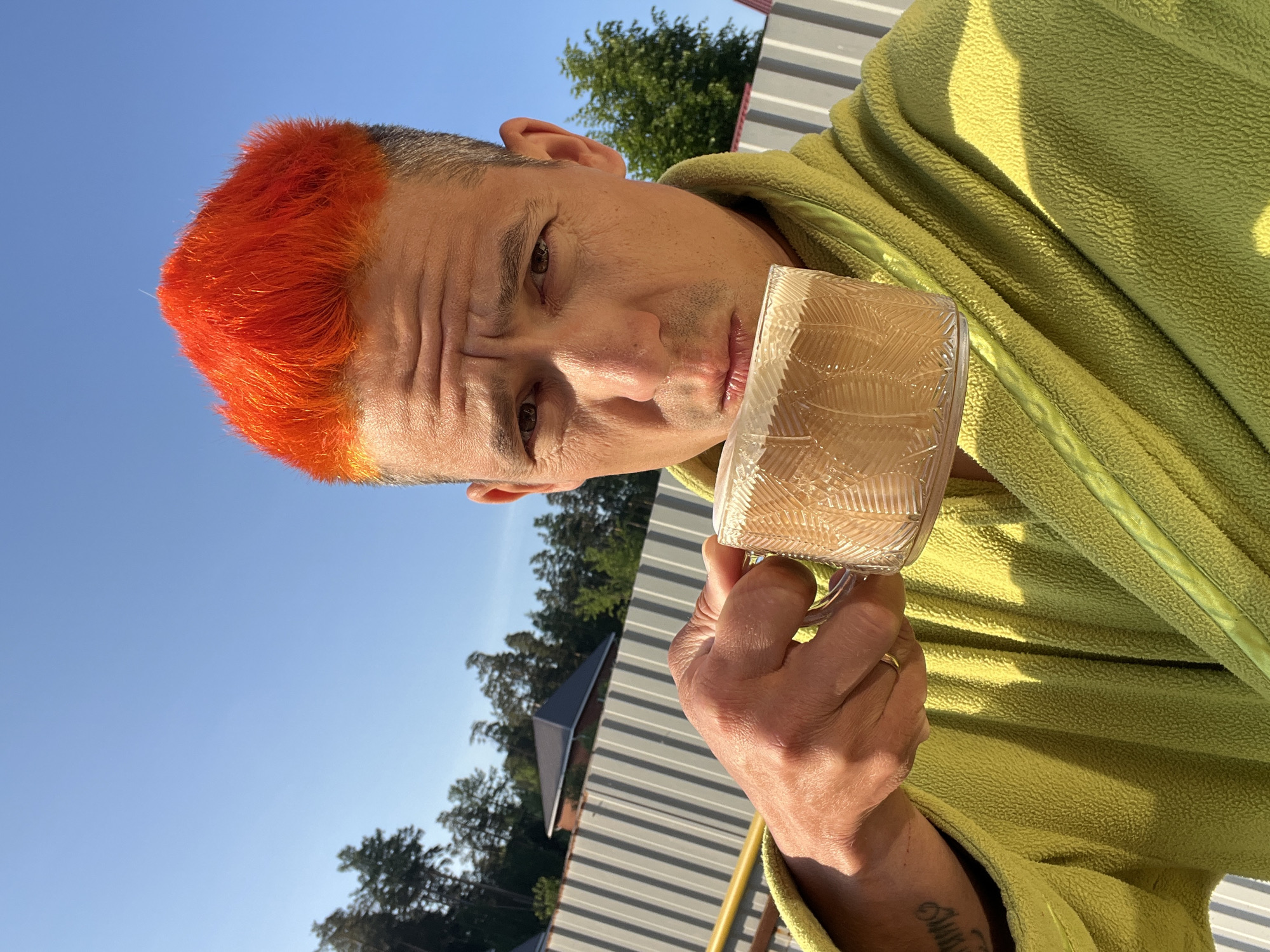 A man with short, dyed orange hair, wearing a light green fleece robe, is holding a clear glass mug, likely containing a beverage. He is looking at the viewer with a slight frown. A gold ring is visible on his left ring finger. A tattoo is visible on his left wrist. The background includes a clear blue sky, treetops, and a corrugated metal fence. It is daytime. While I can see the image, I am unable to pinpoint the specific location within Chaikovsky, Russia.