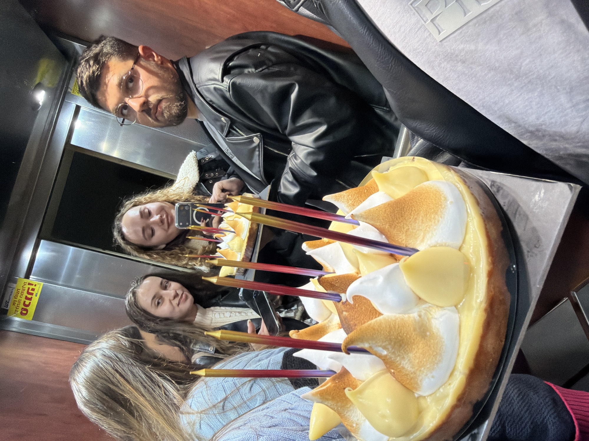 A group of four people are gathered around a cake with candles in what appears to be an elevator in Ramat Gan, Israel.
The individuals consist of three young women and a man. One woman is holding a phone and taking a photo. The man is wearing glasses and a black leather jacket. The women are wearing casual clothing.
The cake is adorned with white and yellow meringue and lit with colorful candles.
The environment is an elevator interior, with metallic walls and a yellow sign visible in the background, suggesting a location within the city of Ramat Gan, Israel. The scene suggests a birthday celebration or a similar celebratory occasion.