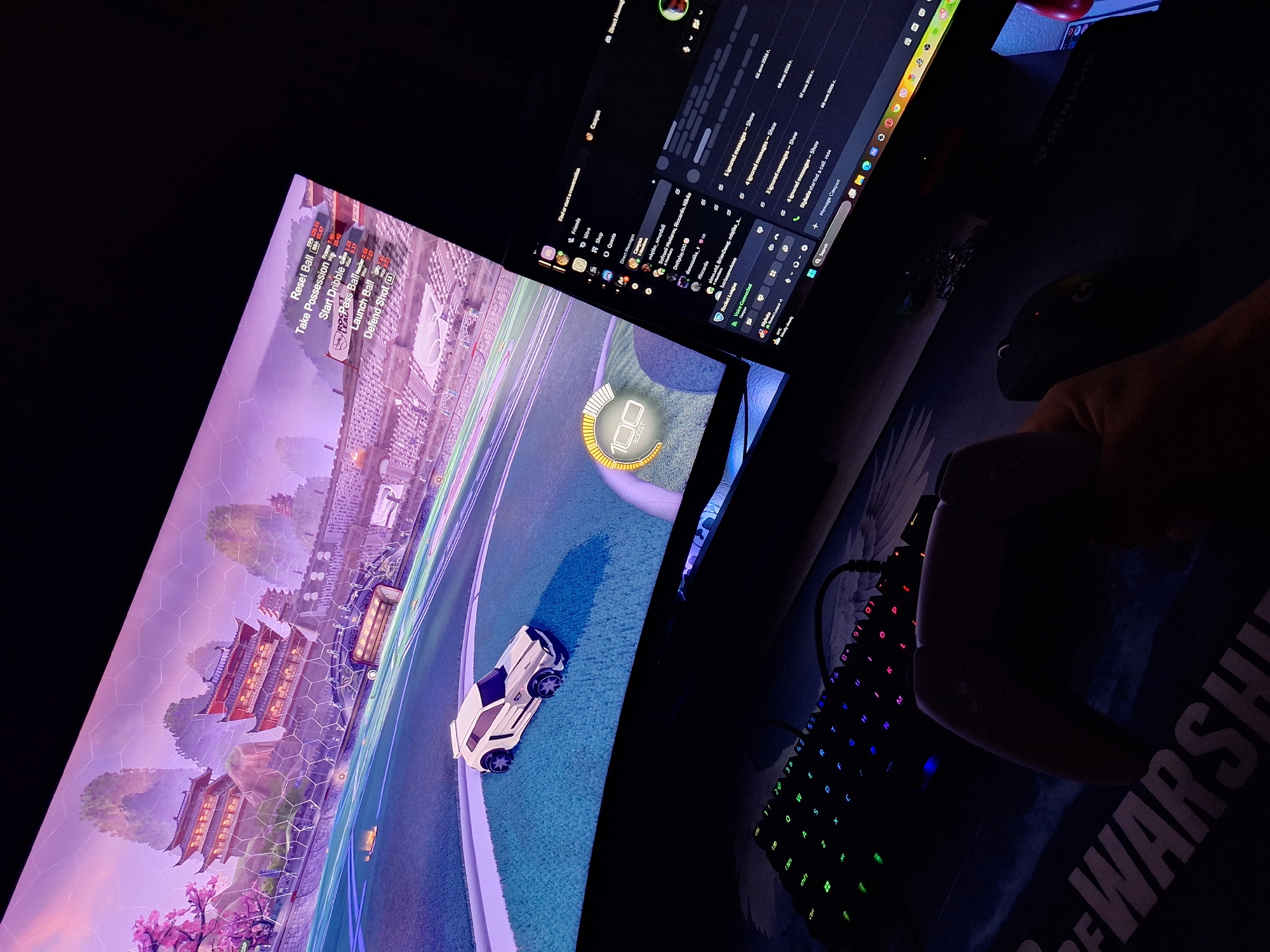 The image shows a person playing a video game called Rocket League on a curved monitor. The game displays a colorful arena with a white car and a boost meter indicating "100". To the right of the monitor is another screen, possibly a laptop, displaying a Discord chat interface. In the foreground, a gaming keyboard with RGB lighting and a game controller are visible. The setting appears to be a dimly lit room. There is no indication of the location being in Varna, Bulgaria.