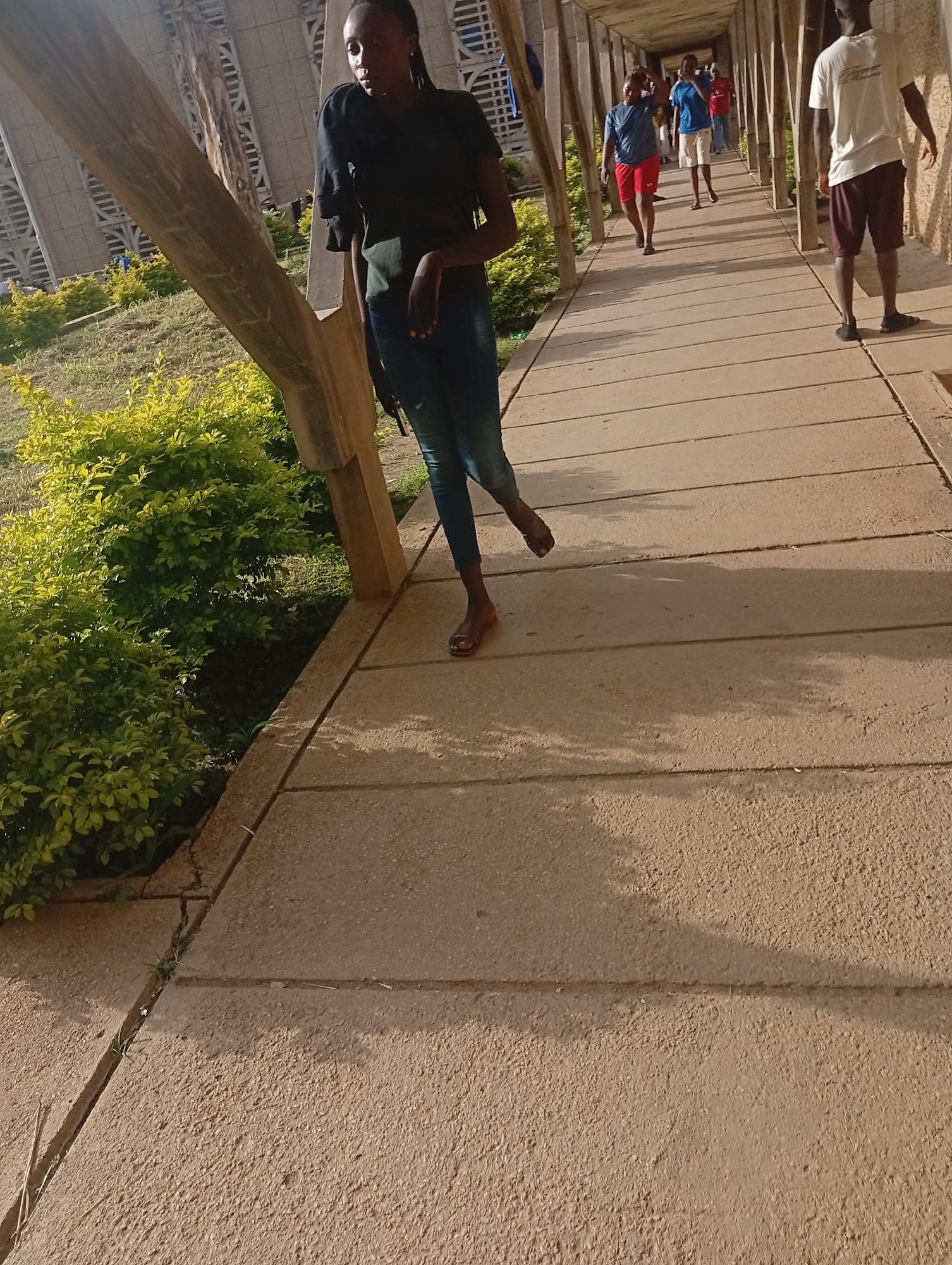 In an outdoor, covered walkway, a young woman of African descent with braided hair, wearing a dark t-shirt and blue jeans, stands on a concrete path. She is facing left, with her right leg extended and bent at the knee, and her left leg is positioned behind her. Her right arm is bent, with her hand on her hip. Sunlight casts shadows across the walkway. Several other individuals, also of African descent, are visible further down the walkway, walking away from the camera. They are wearing casual clothing, including shorts and t-shirts in various colors. The walkway is supported by wooden beams and is lined with green foliage on the left side. The background shows a portion of a large, angular building with a patterned facade. The overall environment appears to be a public space, possibly on a college campus or similar institution.