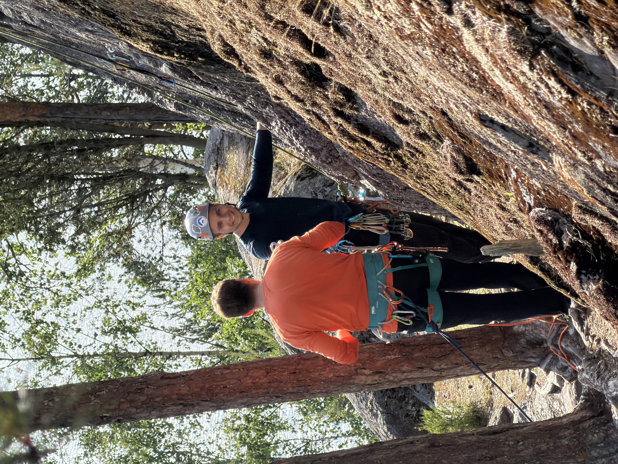Two individuals are engaged in rock climbing near a body of water, likely a lake, in Ruch'i, Russia. The person in the foreground, wearing an orange long-sleeved shirt and black pants with a teal harness, is facing away from the camera, towards the rock face. They are equipped with climbing gear, including quickdraws attached to their harness. The second individual, in the middle ground, is wearing a dark long-sleeved shirt and a helmet with a patch, and is smiling at the camera, looking towards the rock. They are also wearing a harness and carrying climbing equipment. The rock face is covered in moss and vegetation, indicating a natural outdoor environment. Several trees are visible in the background, with the shimmering surface of water behind them. The ground is rocky and covered with pine needles and small bushes. The scene appears to be a sunny day, as indicated by the lighting and shadows.