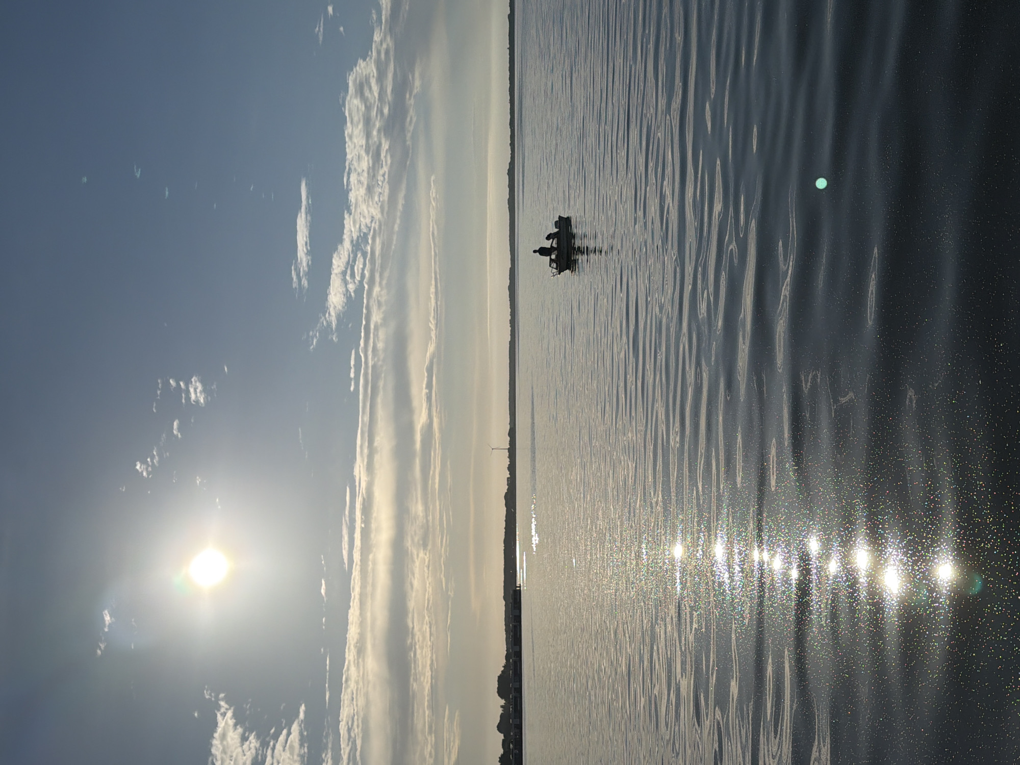 The media displays a wide shot of a body of water with a person or people in a small boat. The sun is visible in the sky, casting a bright reflection on the water's surface. The sky is partly cloudy, with the sun illuminating the clouds from behind. The water appears calm, with small ripples and a shimmering effect from the sunlight. The overall scene evokes a sense of peace and tranquility. The image is oriented vertically, with the sky on the left and the water on the right. A distant shoreline is visible, with trees and a wind turbine, suggesting a rural or coastal location. The time of day appears to be morning or late afternoon, given the angle of the sun. The weather seems clear and pleasant. No text is visible in the image. The location context provided is Ribnitz-Damgarten, Germany.