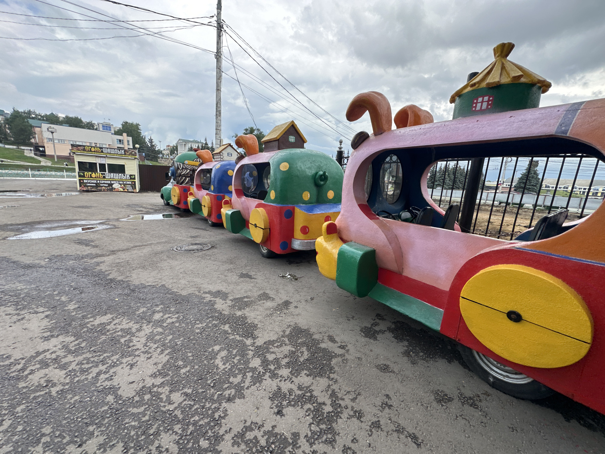 In Saransk, Russia, a row of colorful, toy-like train cars, likely a children's amusement ride, is parked on a paved surface. The train cars are brightly painted in red, green, pink, and yellow, with whimsical features such as cartoonish ears on the front car. The first car has an orange roof that resembles a stylized house. Behind the train, a small kiosk with a sign reading "Огонь-Шашлык" (Fire-Shashlik) suggests a food vendor. Further in the background, buildings and trees are visible under a cloudy sky, indicating an outdoor urban setting. The ground is asphalt, wet in places, with puddles reflecting the overcast sky and surrounding elements. The overall scene depicts a temporary outdoor amusement area within the city.
