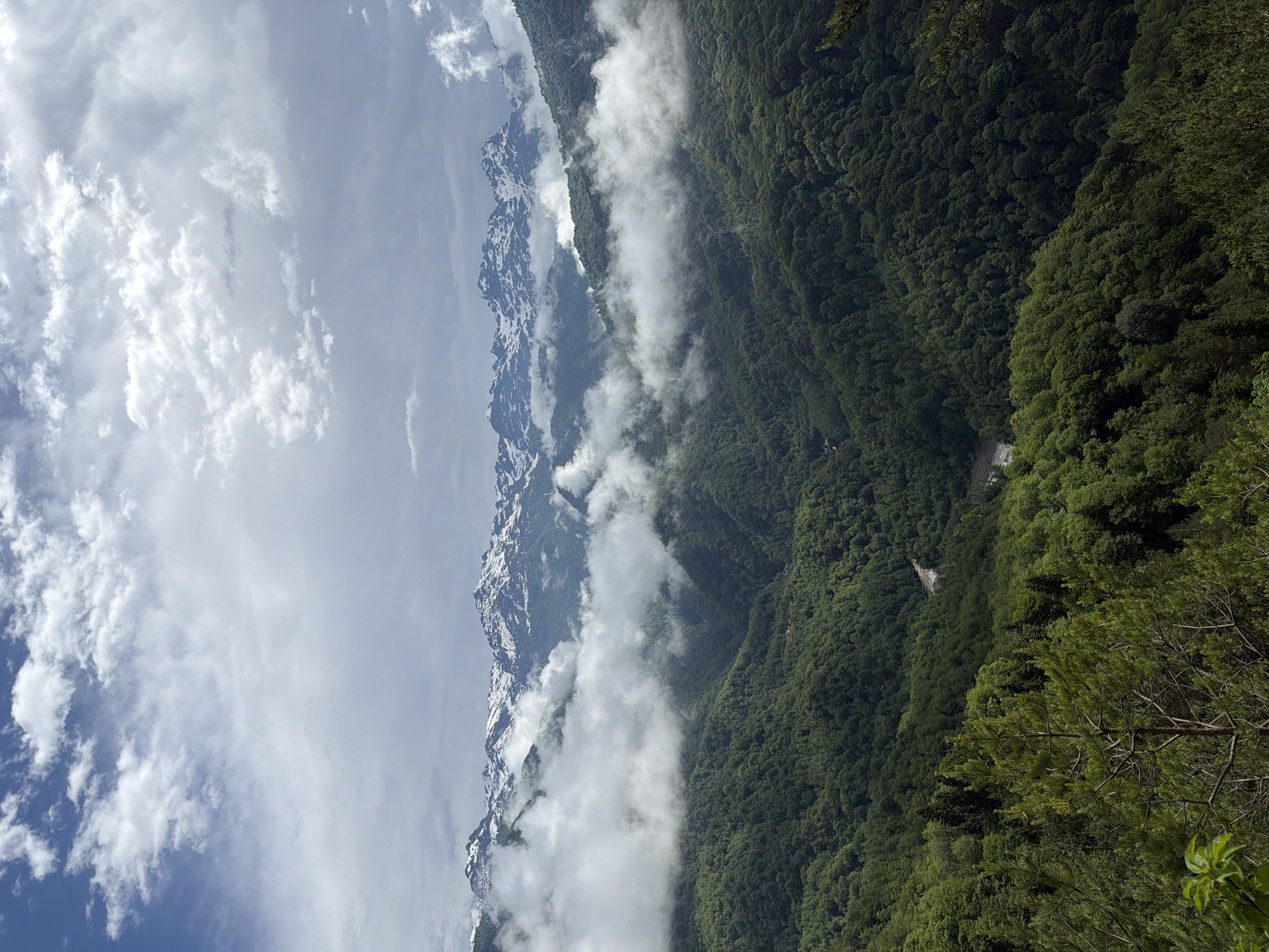 This is a vertical, high-angle shot of a mountainous, forested landscape under a cloudy sky. The foreground features the tops of tall evergreen trees. The midground is dominated by rolling, densely forested green hills, with a visible winding road and a small building nestled within the trees. Wisps of white clouds weave through the valleys between the hills. In the background, snow-capped mountains rise majestically, their peaks partially obscured by clouds. The sky is a mix of light blue and gray clouds, with some brighter patches indicating sunlight filtering through. The scene appears to be a natural, undeveloped area, characteristic of the mountainous terrain around Potskho-Etseri, Georgia. There are no people or discernible human activity visible in the image.