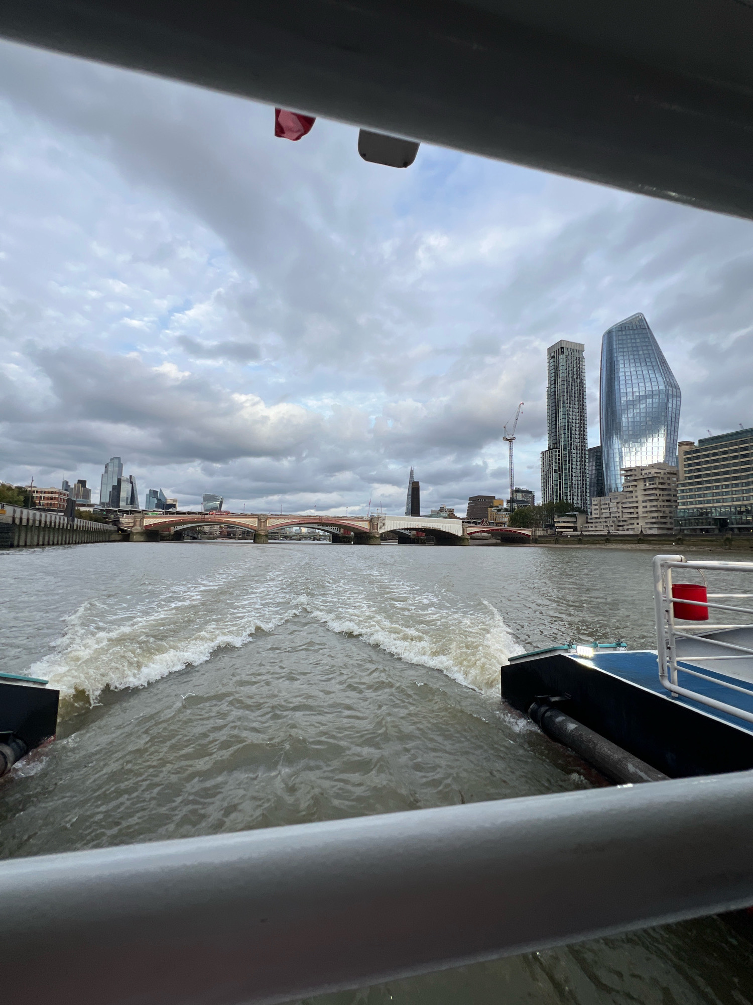 The image captures a view from a boat on the River Thames in London, United Kingdom. The foreground shows the wake of the boat creating white, choppy waves in the murky water, with a portion of the boat's railing and hull visible on the right and left sides, respectively. In the mid-ground, a red-bricked bridge spans the river, with a complex arrangement of support arches and roadway above. Beyond the bridge, the London skyline is dominated by a cluster of modern skyscrapers, including a tall, slender building with a patterned facade and a distinctive, glass-covered, uniquely shaped tower. In the background, the sky is heavily overcast with dark, dramatic clouds. A small red flag is visible near the top edge of the image, attached to an unseen structure.